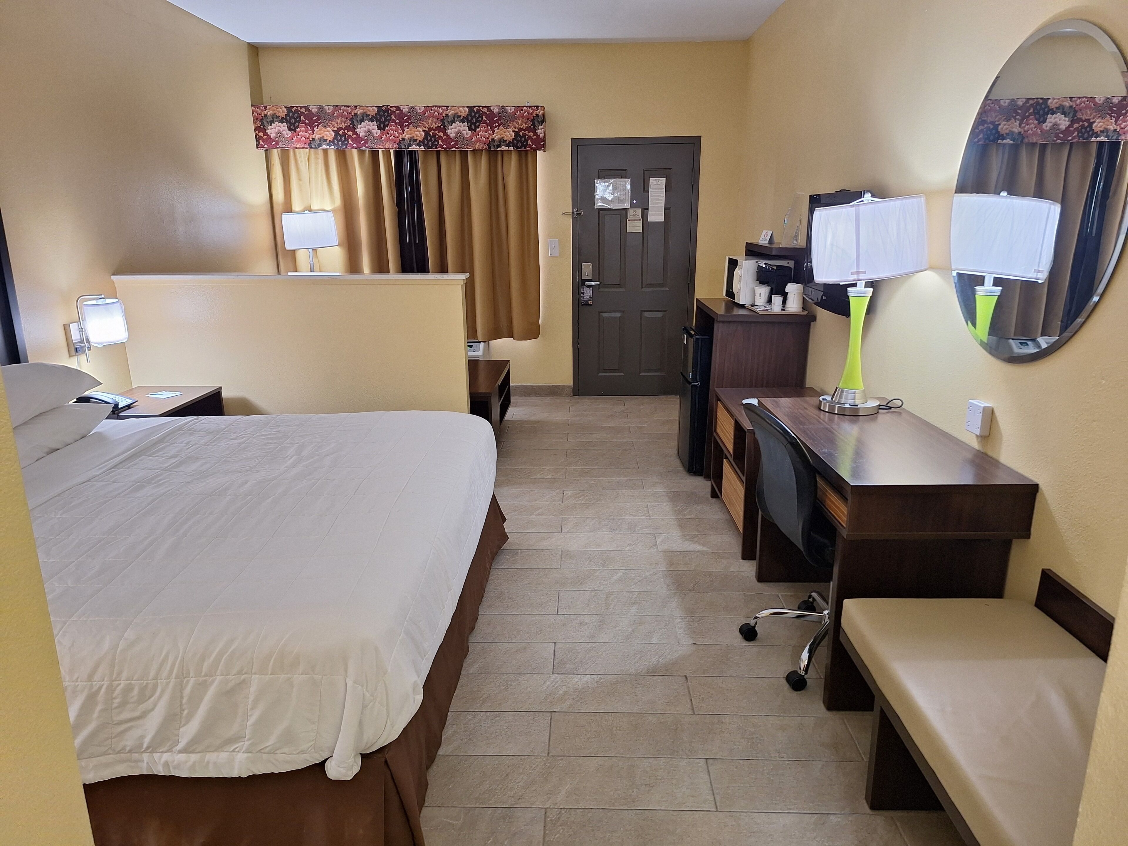 Habitación de hotel con cama, escritorio, silla, espejo, puerta y una pequeña kitchenette.