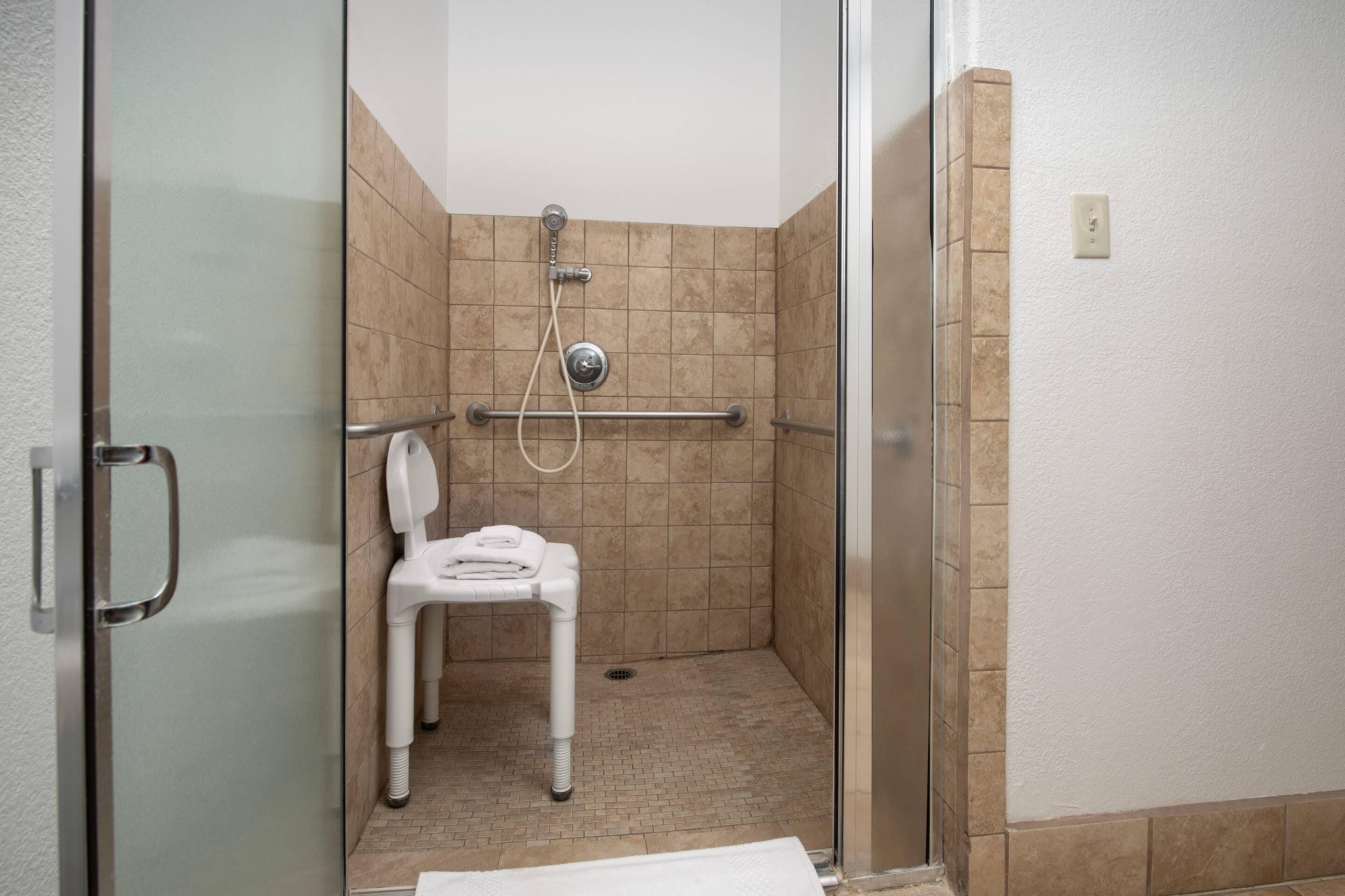 A glass-enclosed shower stall with a white chair, a handheld showerhead, and a wall-mounted control panel.