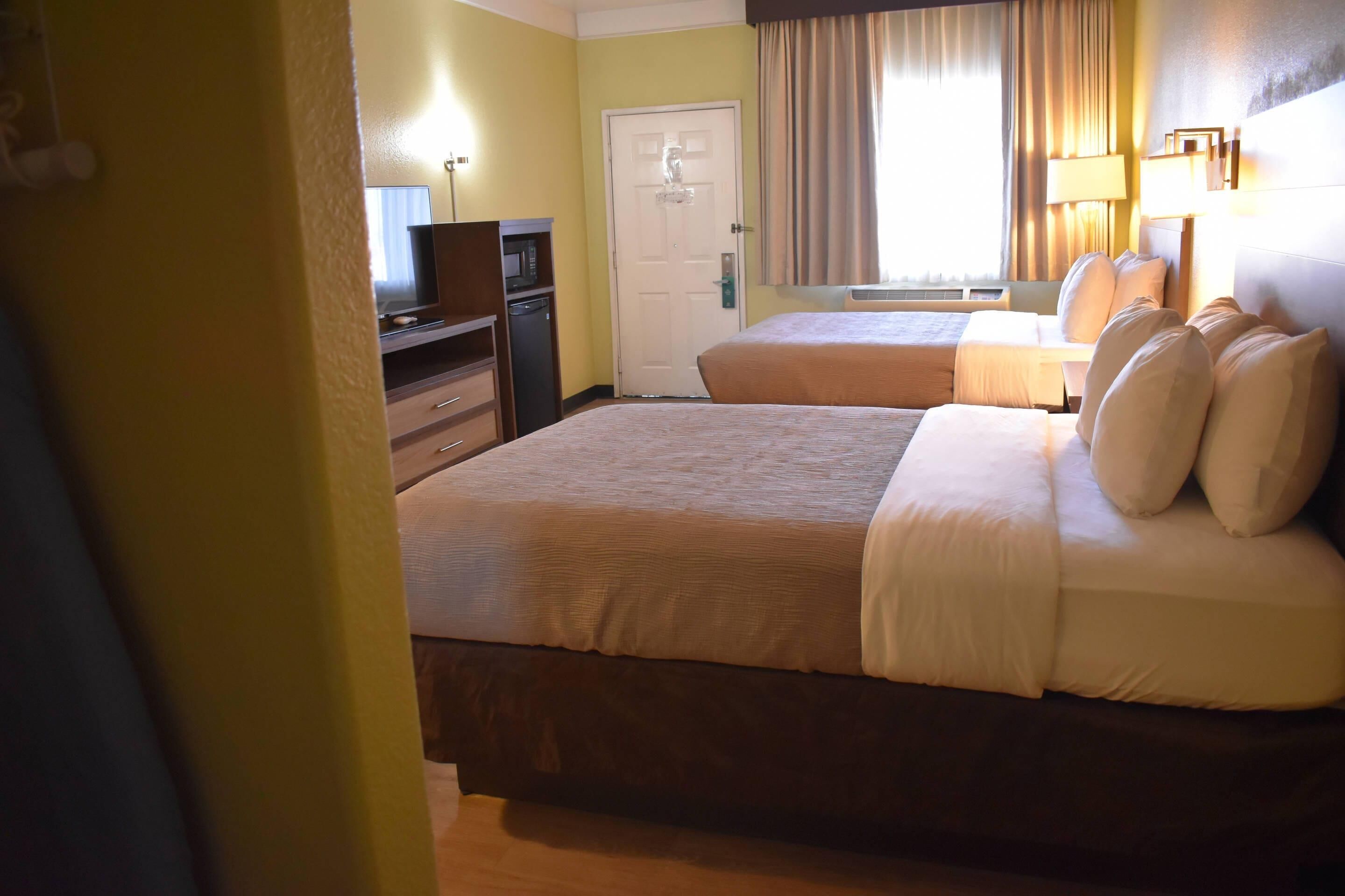 A hotel room with two beds, a desk with a computer, a door, and a window with curtains.