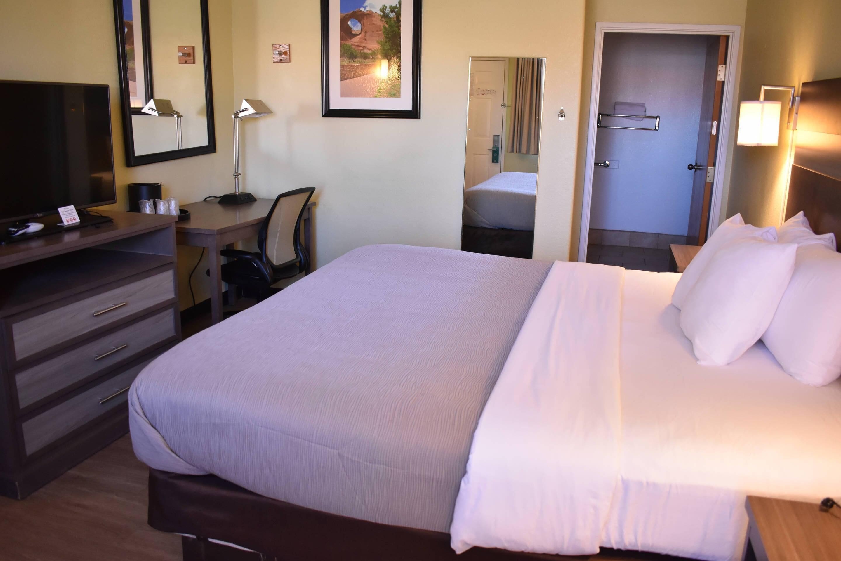 Una camera d'albergo con un letto, una scrivania con un computer, una televisione, uno specchio e una porta che dà sul bagno.