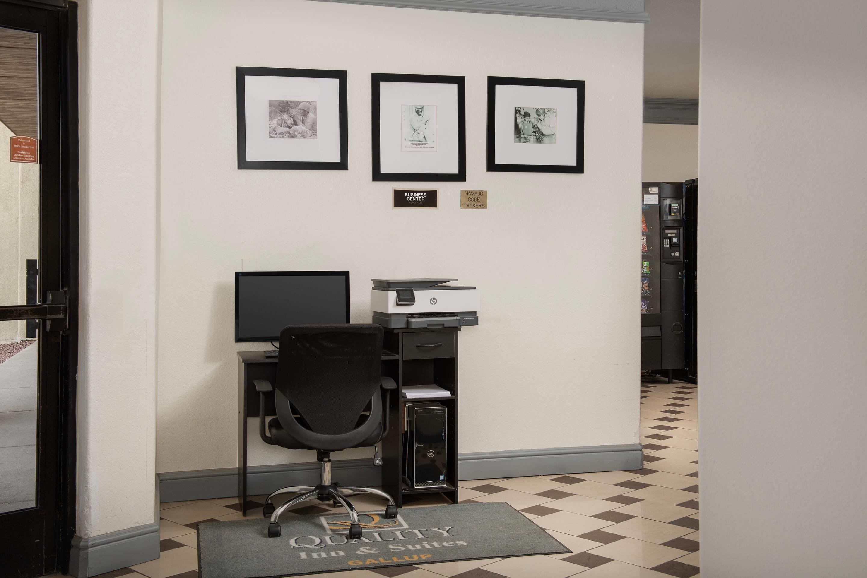 A hotel office area with a desk, chair, computer, printer, and framed pictures on the wall.