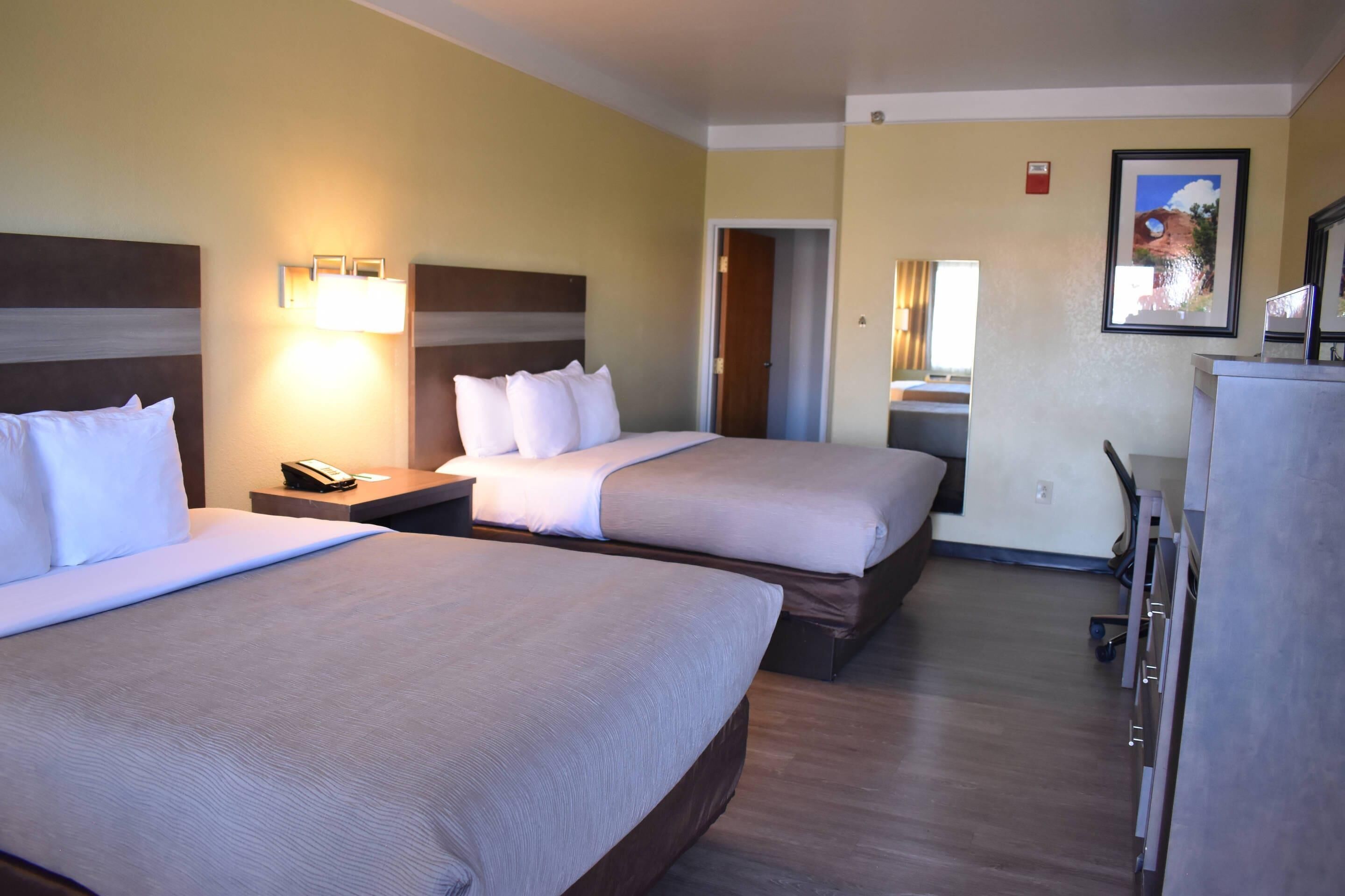 A hotel room with two beds, a desk, a chair, a lamp, and a framed picture on the wall.
