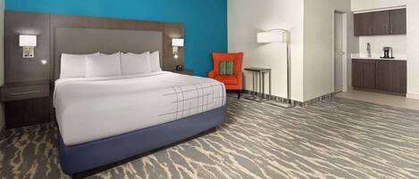 A hotel room with a large bed, a nightstand, a chair, a desk, and a kitchenette.