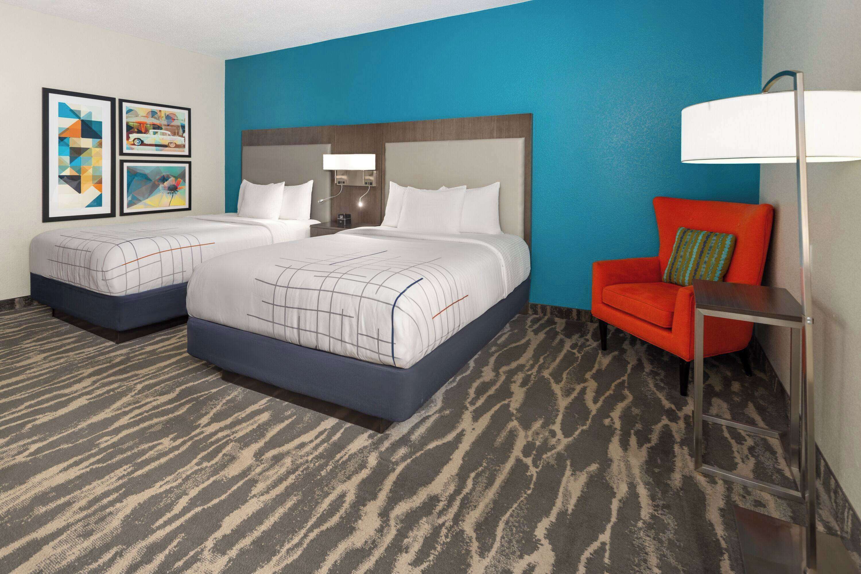 A hotel room with two beds, a red armchair, a standing lamp, and artwork on the wall.