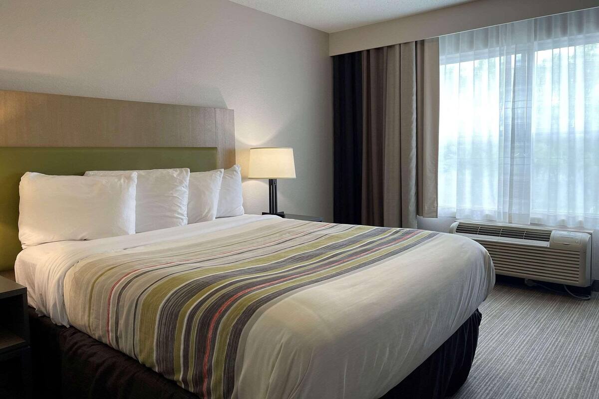 A hotel room with a large bed, a headboard, a bedside table, a lamp, a window with curtains, and an air conditioning unit.