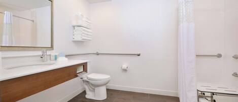 Baño con lavamanos blanco, inodoro y zona de ducha, que incluye una barra de apoyo y un perchero para toallas.