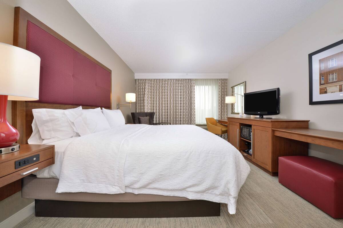 A hotel room with a large bed, a desk with a television, a red bench, and a wall-mounted TV.
