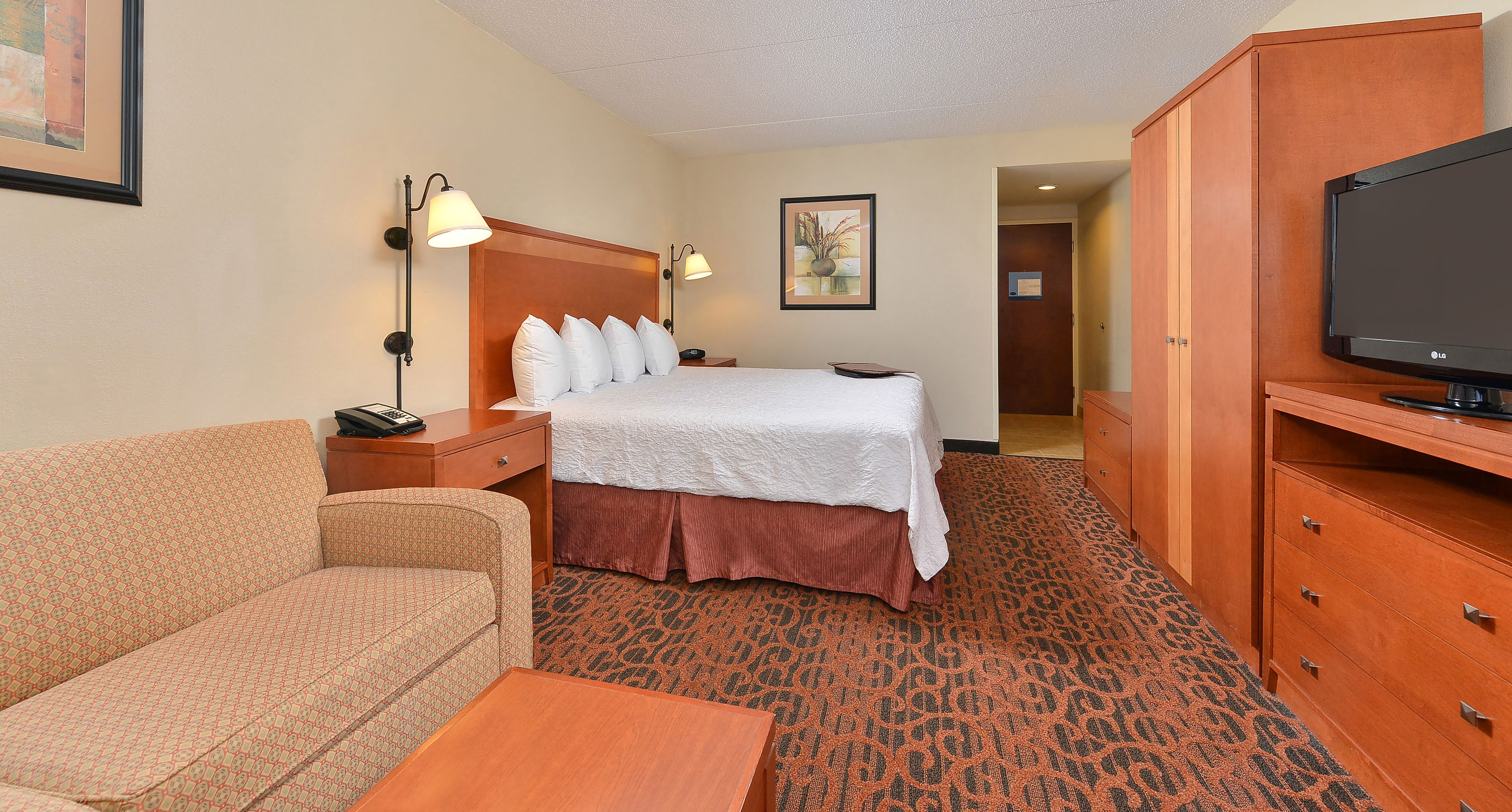 A hotel room with a bed, a TV, a sofa, a nightstand, and a wooden wardrobe.