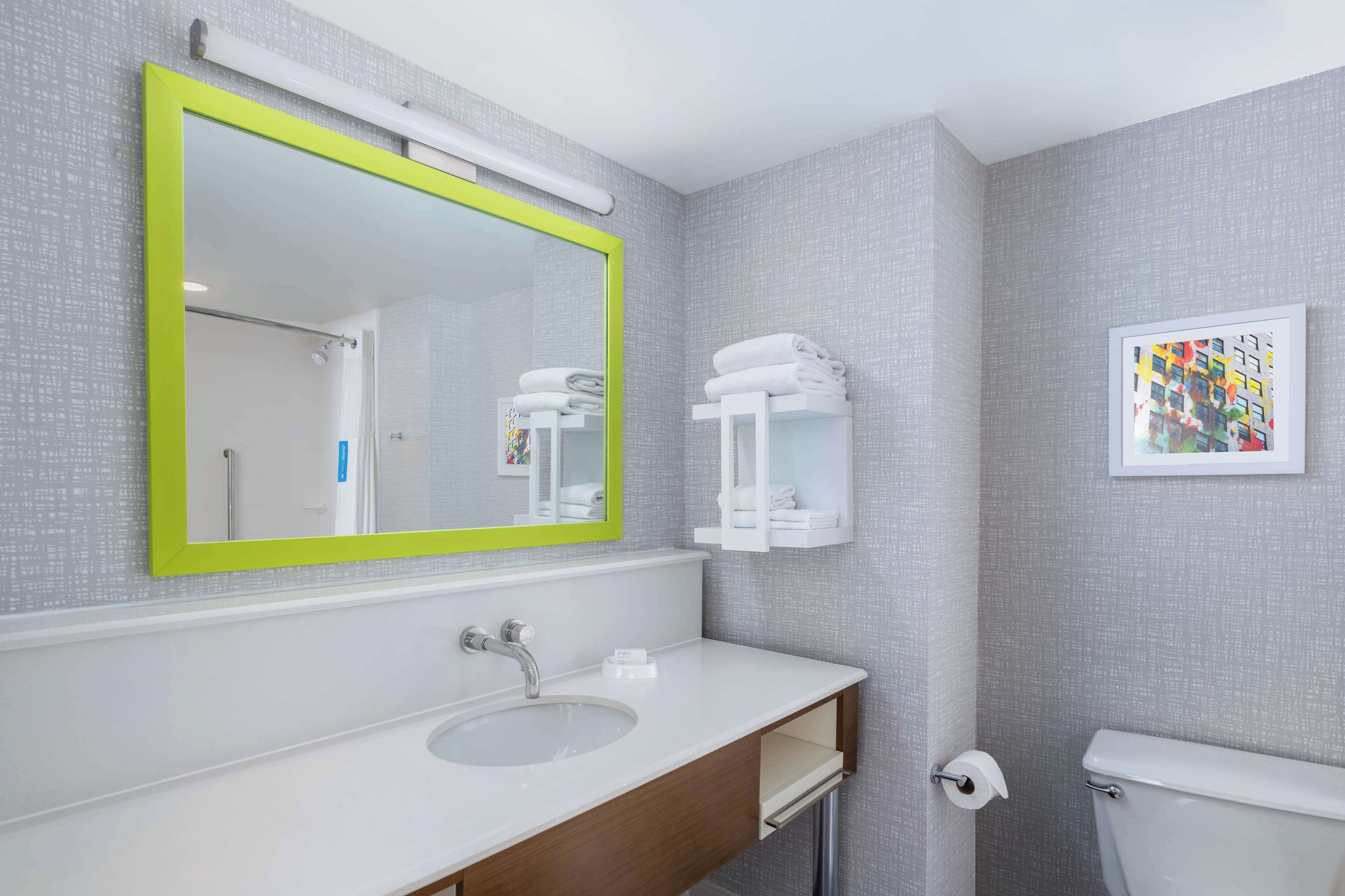 Une salle de bain moderne avec un lavabo blanc, un miroir à cadre vert, des toilettes et une œuvre d’art encadrée accrochée au mur.