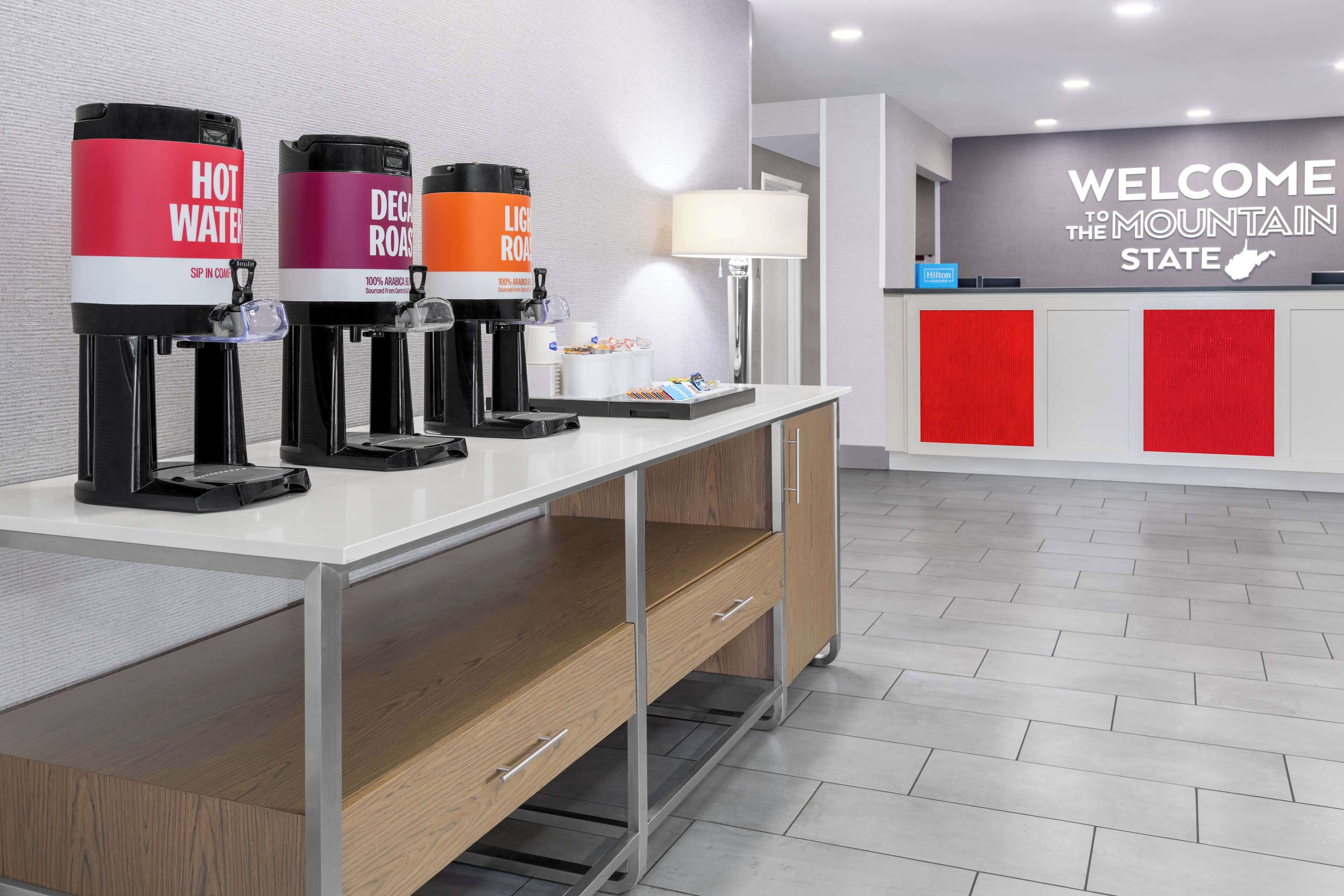 A hotel reception area with a white reception desk, a "Welcome to the Mountain State" sign, and three beverage dispensers labeled "Hot Water," "Decaf Roast," and "Light Roast."