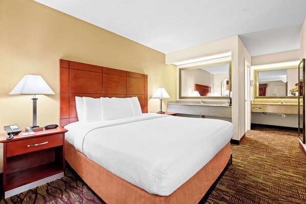 A hotel room with a large bed, a bedside table with a lamp, a mirror, and a bathroom with a sink and mirror. - Best Western Inn (Clanton)