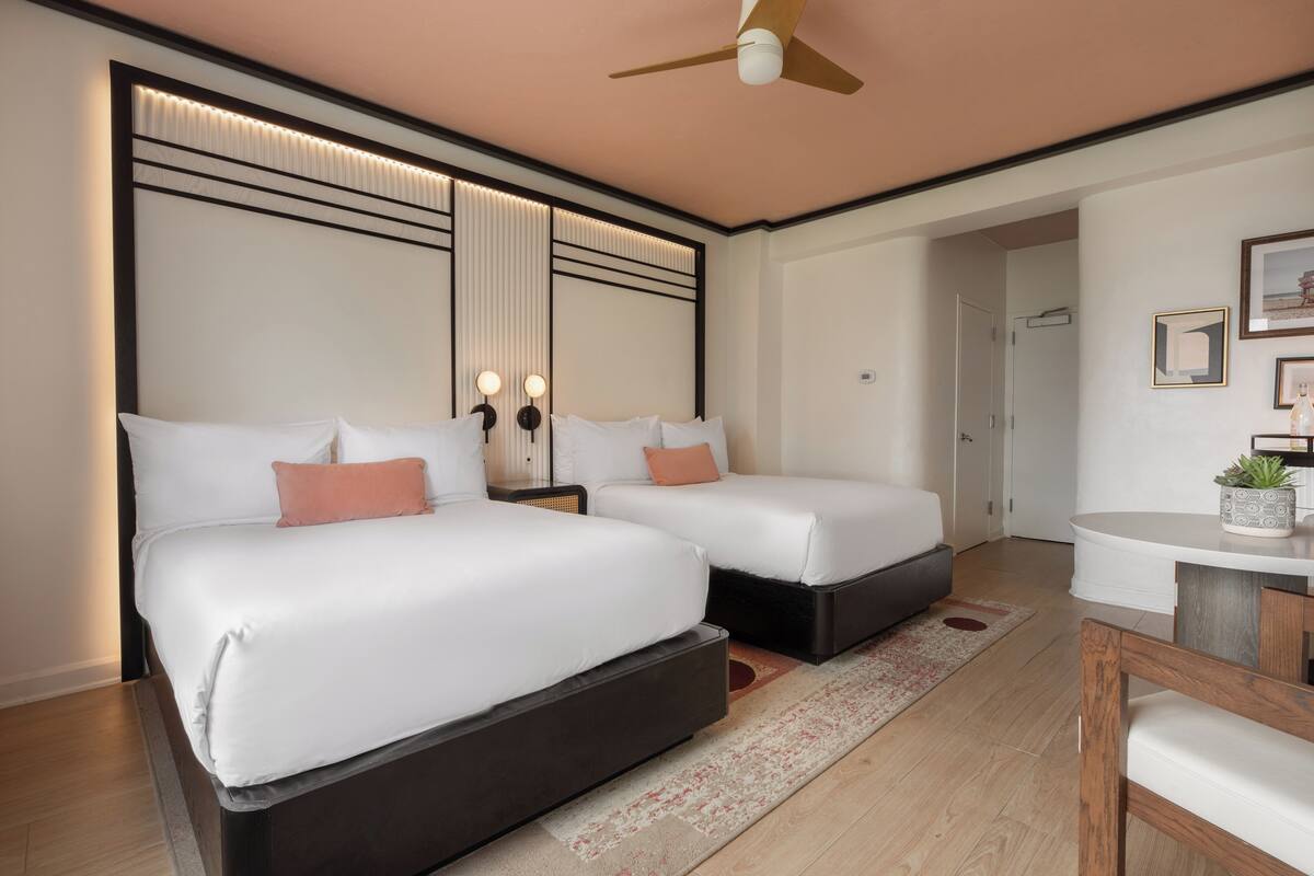 A hotel room with two beds, a ceiling fan, a small table with a plant, and a door leading to another room.