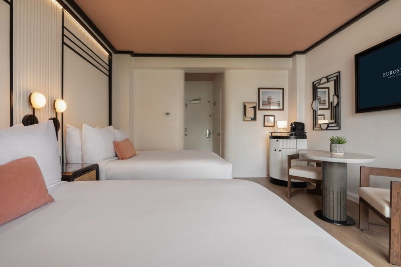 A hotel room with two beds, a desk, a chair, and a small table with a plant.