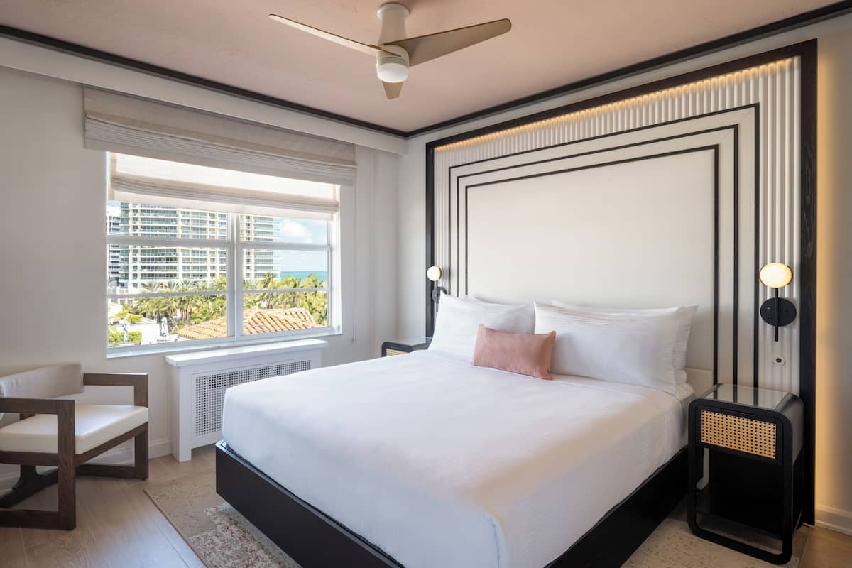 A modern hotel room with a large bed, a chair, a nightstand, and a view of the cityscape through the window.