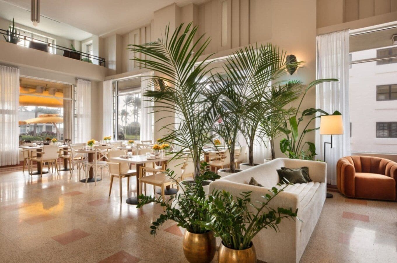 A spacious hotel lobby with a high ceiling, multiple dining tables, comfortable seating, and large potted plants.