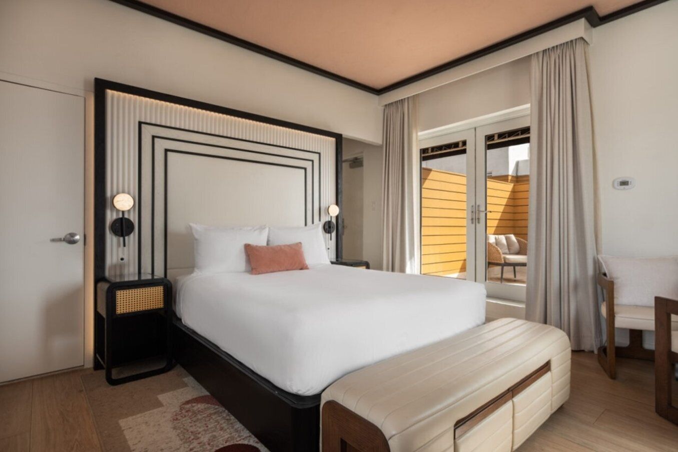 A hotel room with a large bed, a bedside table, a bench, and a view of the outside.
