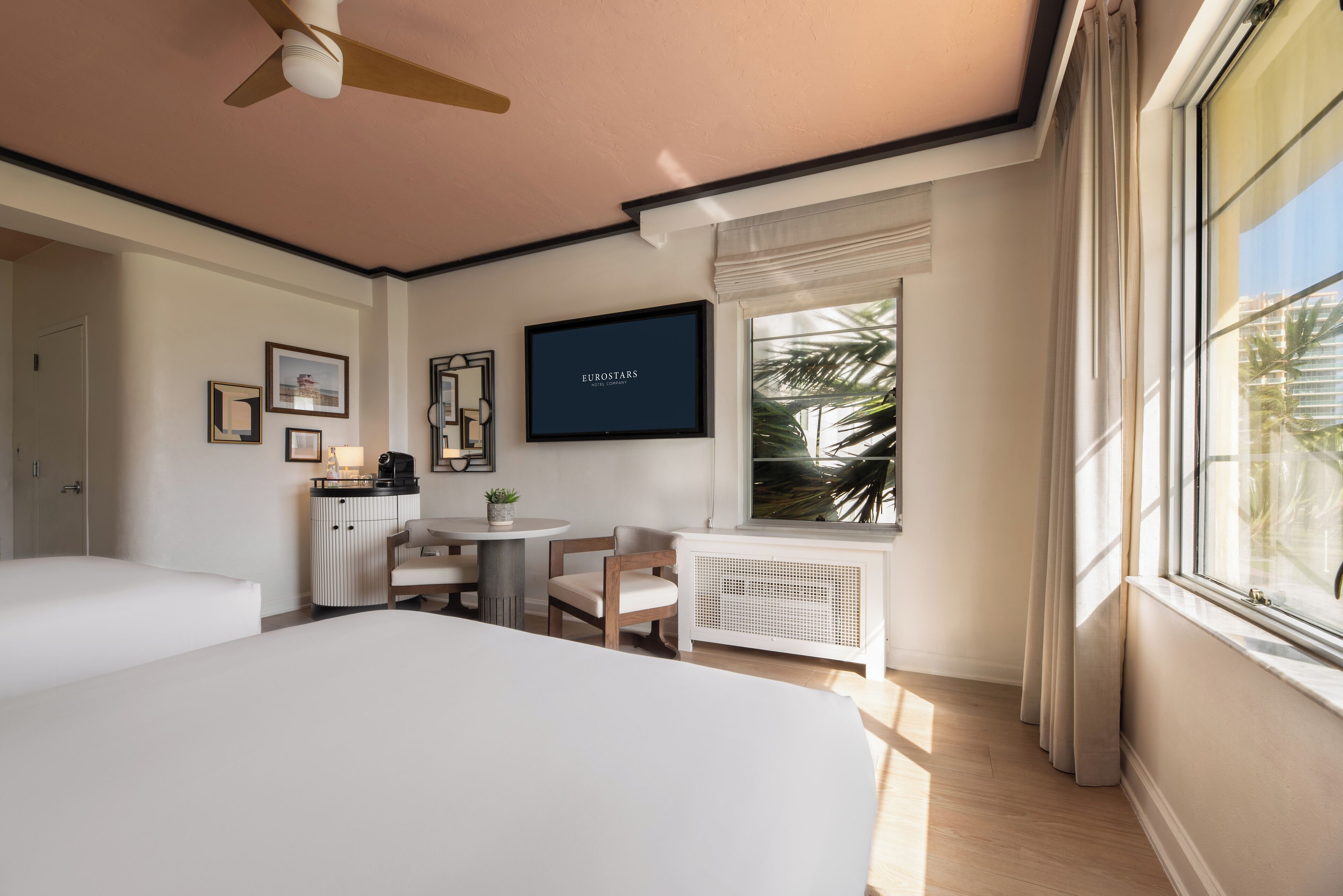 A hotel room with a bed, a small table with chairs, a TV on the wall, and a window with curtains.