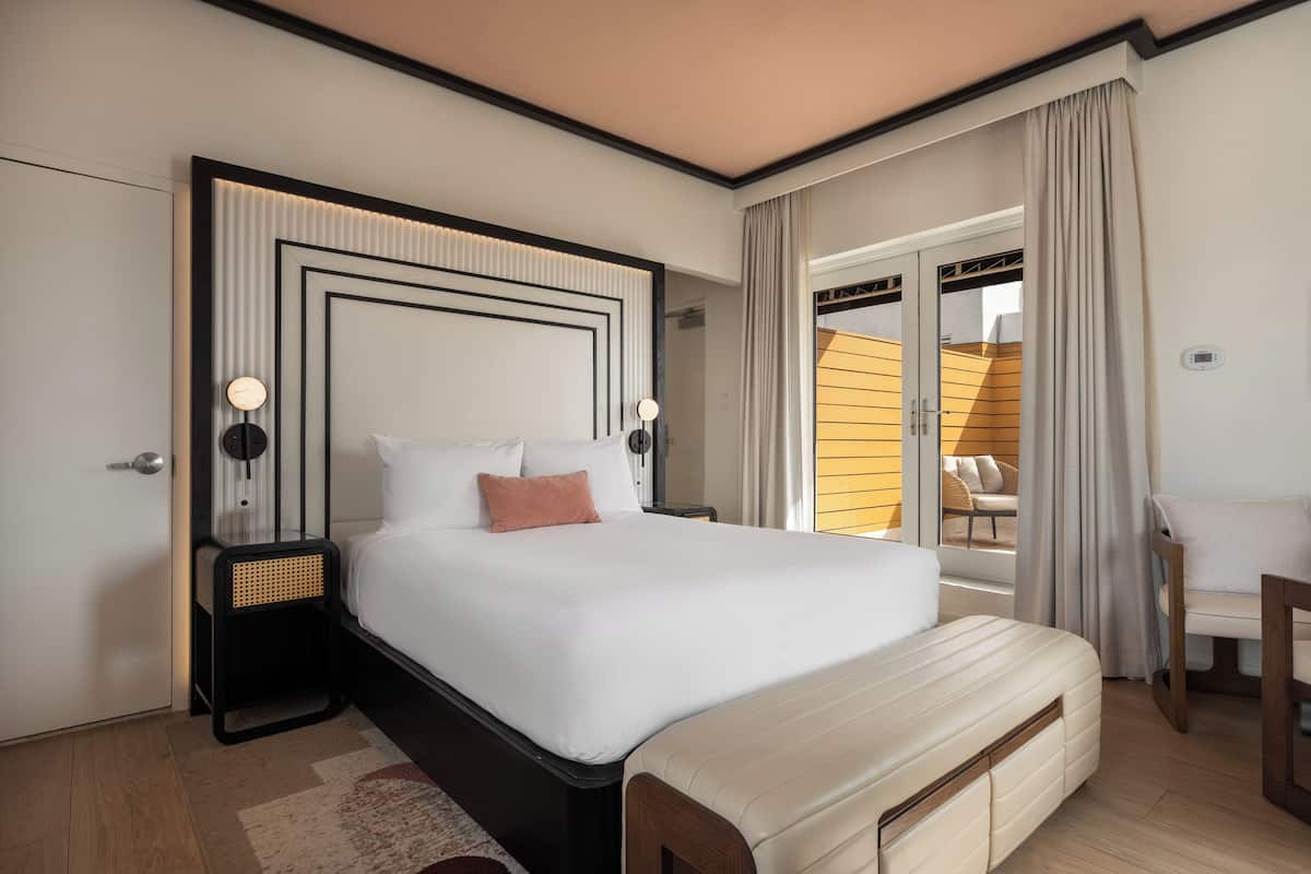 A modern hotel room with a large bed, a bedside table, a bench, and a view of the outdoor seating area.