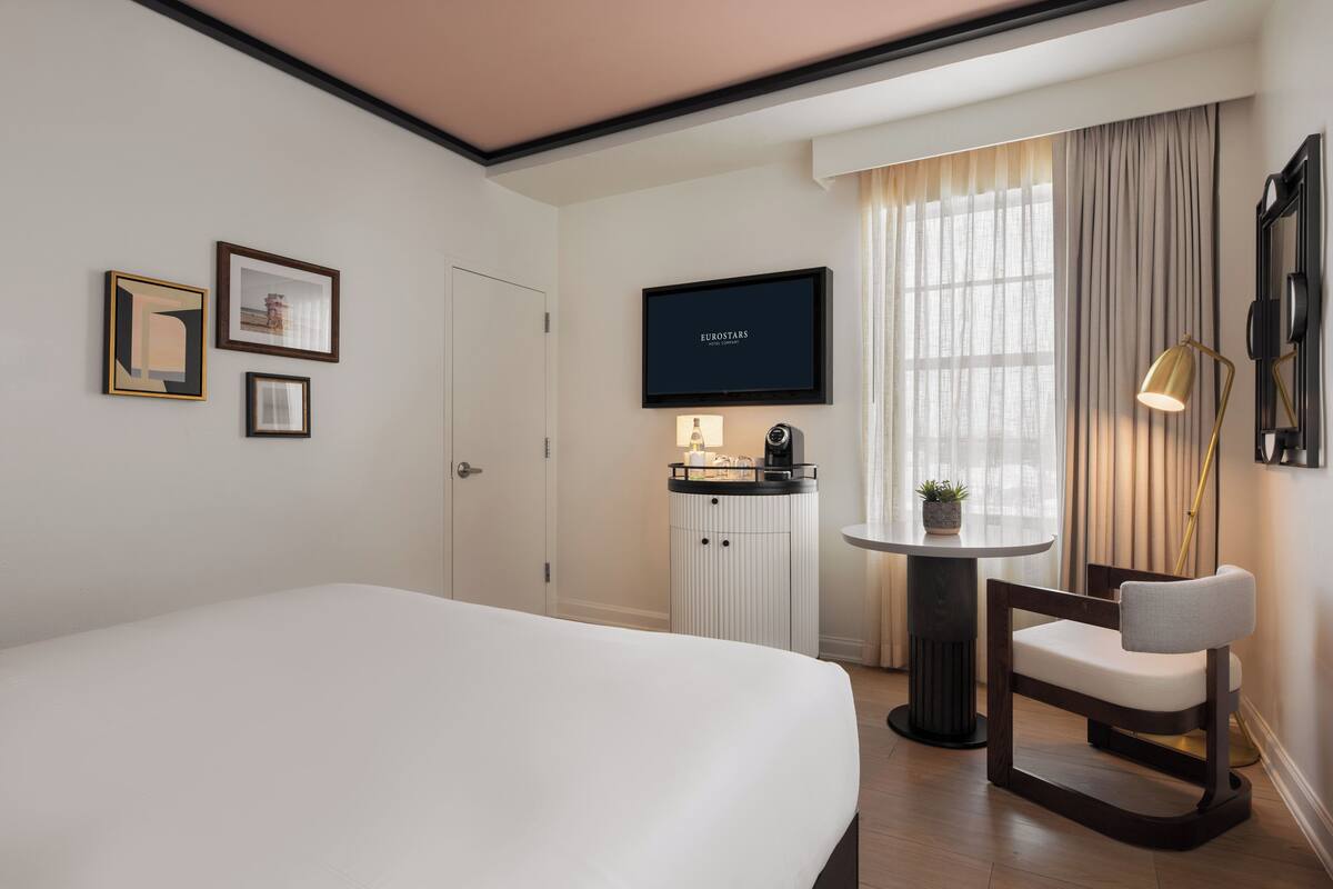 A hotel room with a large bed, a television on the wall, a small table with a lamp, and a chair.