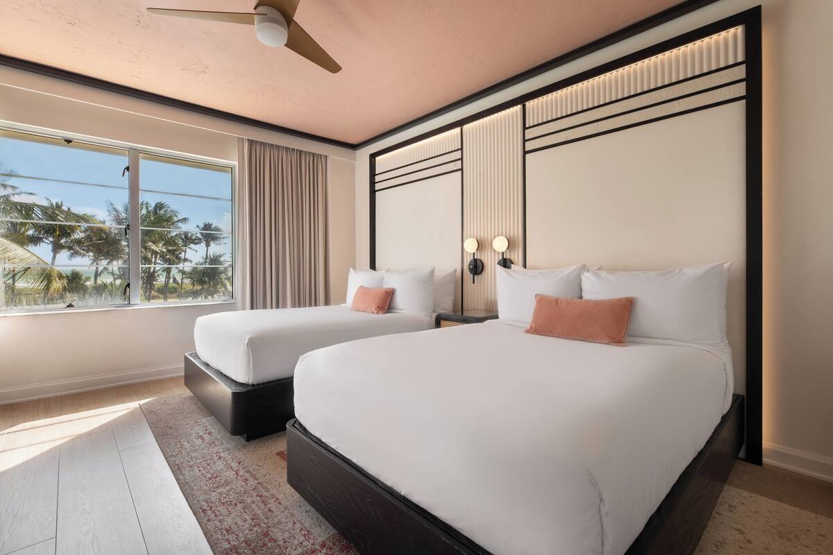 A hotel room with two beds, a large window with a view of palm trees, a ceiling fan, and a modern design.