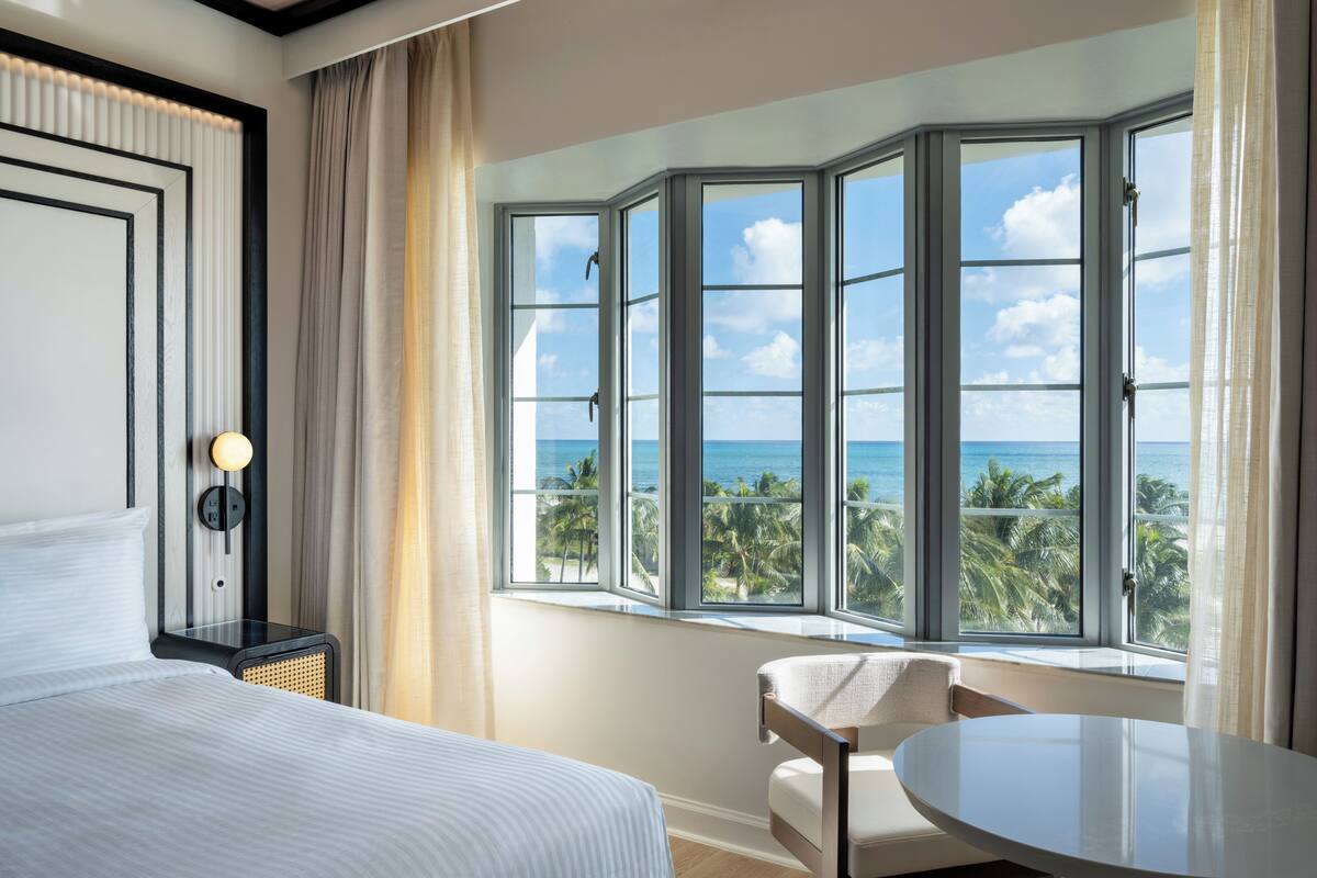 A hotel room with a large bay window offering a view of the ocean and palm trees.