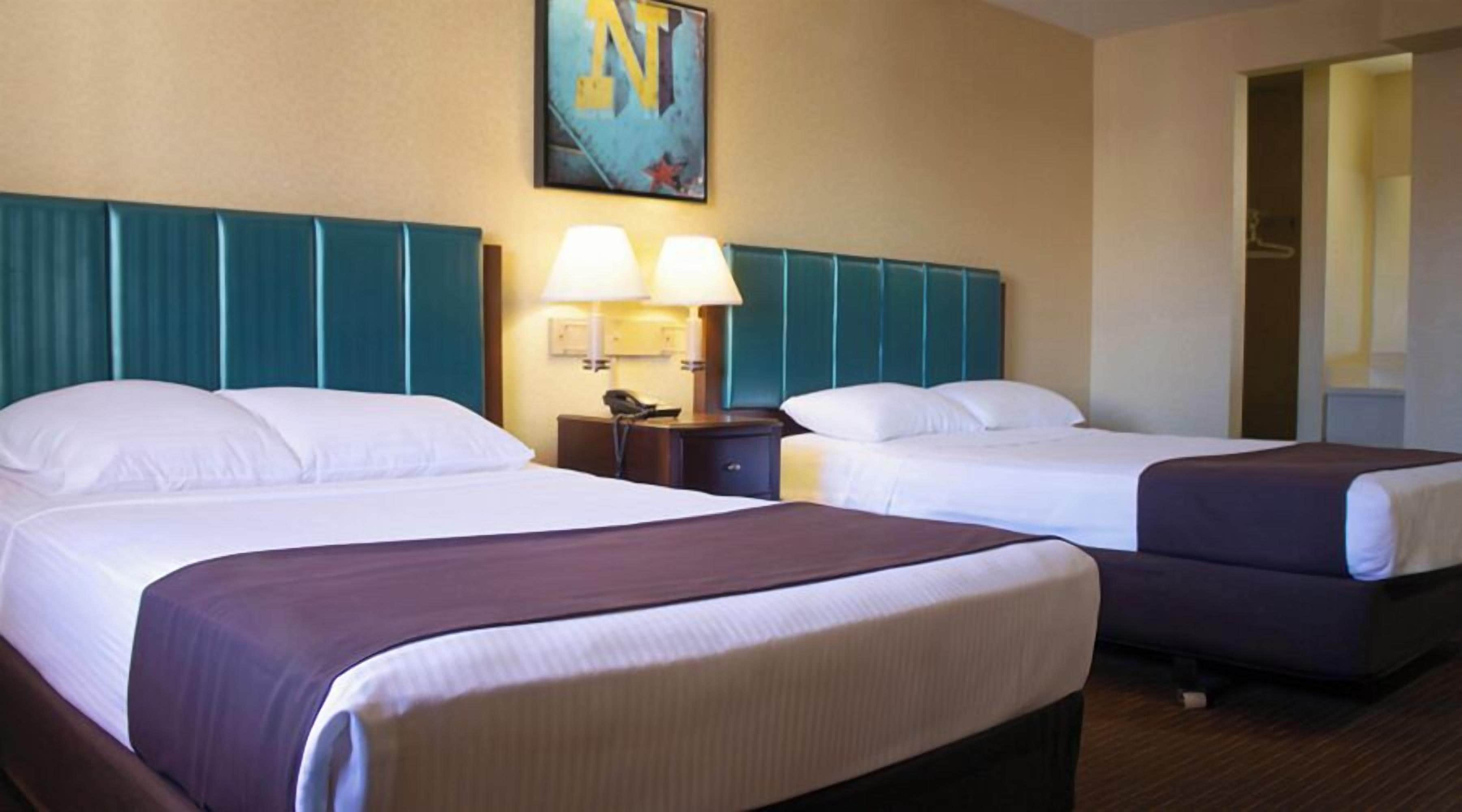 A hotel room with two beds, a headboard with teal panels, a painting on the wall, and a nightstand with a lamp.