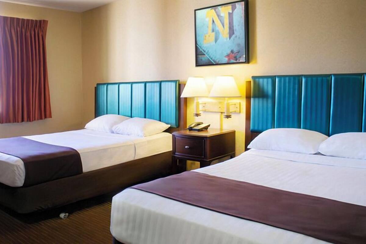A hotel room with two beds, a window with curtains, a wall-mounted painting, and a nightstand with a lamp.