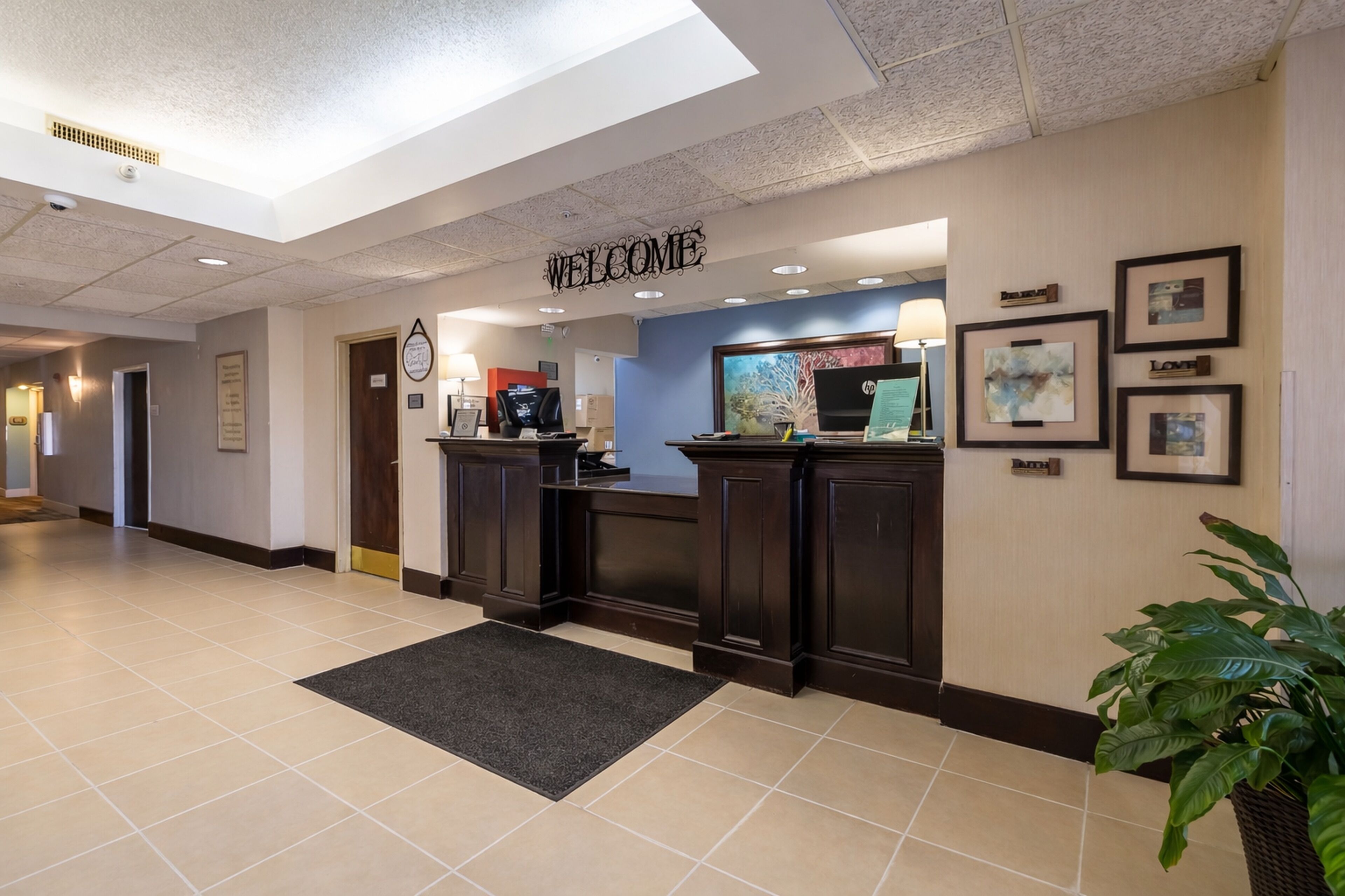 Zona de recepción de hotel con un cartel de 'WELCOME', un mostrador de recepción, una máquina de café, obras de arte enmarcadas y una planta en maceta.