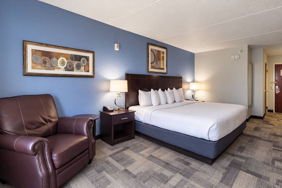 Une chambre d’hôtel avec un grand lit, un fauteuil en cuir marron, deux tables de chevet avec des lampes et deux œuvres d’art encadrées accrochées aux murs.