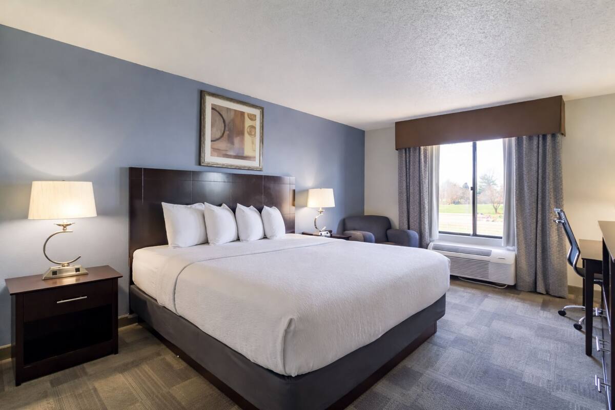Une chambre d’hôtel avec un grand lit, deux tables de chevet avec des lampes, un bureau avec une chaise, une fenêtre avec des rideaux et un tableau au-dessus du lit.
