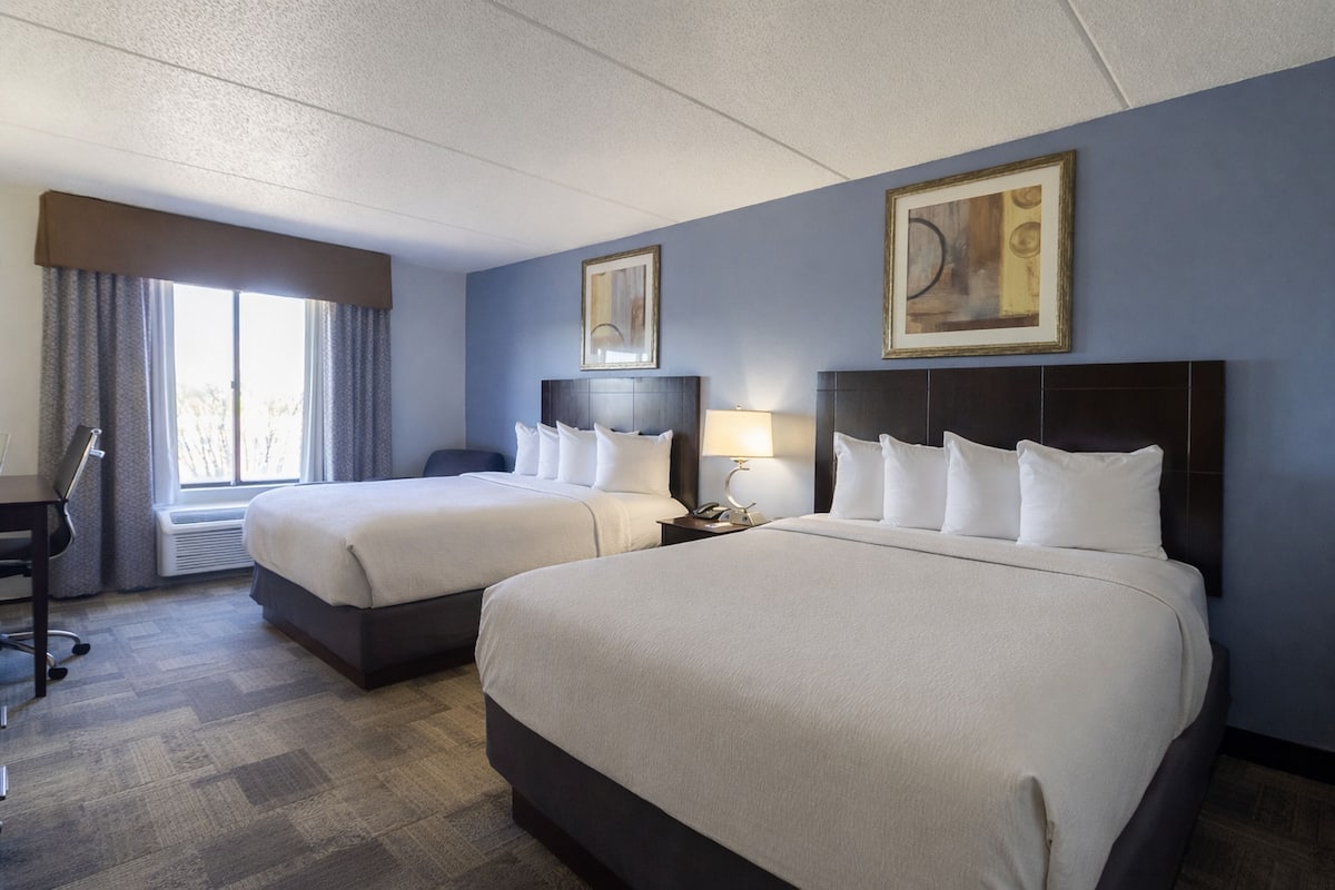 Une chambre d’hôtel avec deux lits, un bureau, une chaise, une lampe et des œuvres d’art accrochées aux murs.
