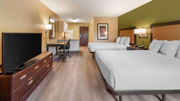 A hotel room with two beds, a desk with a chair, a TV, and a small kitchenette.