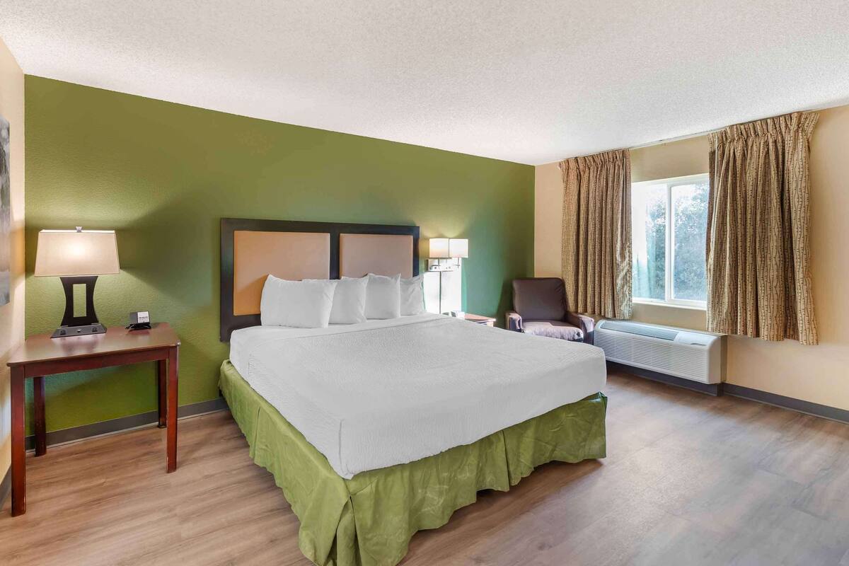 Una habitación de hotel con una cama grande, una mesita de noche con lámpara, una silla y una ventana con cortinas.