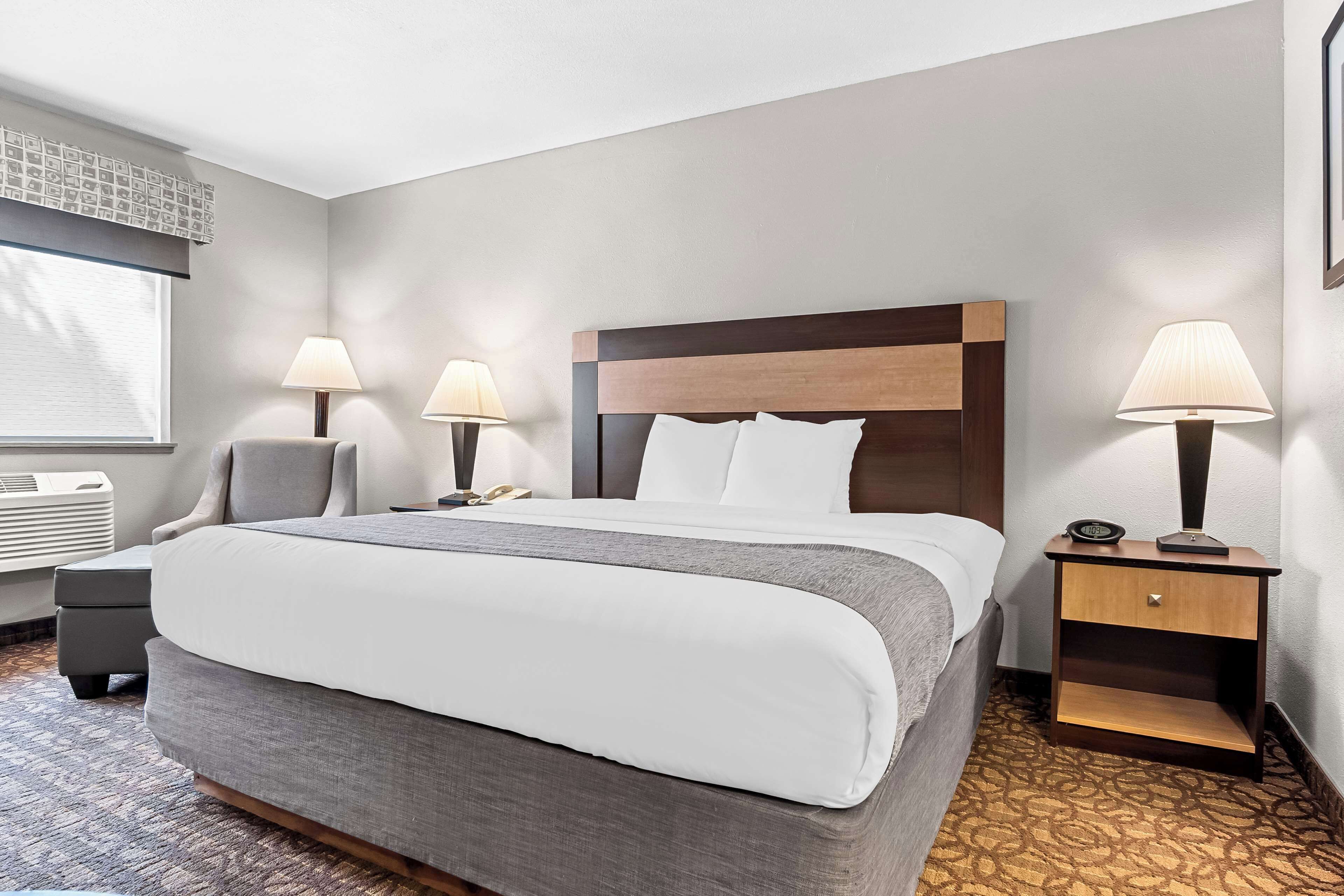 Habitación de hotel con una cama grande, dos mesitas de noche con lámparas, una silla pequeña, un equipo de aire acondicionado y una alfombra estampada.
