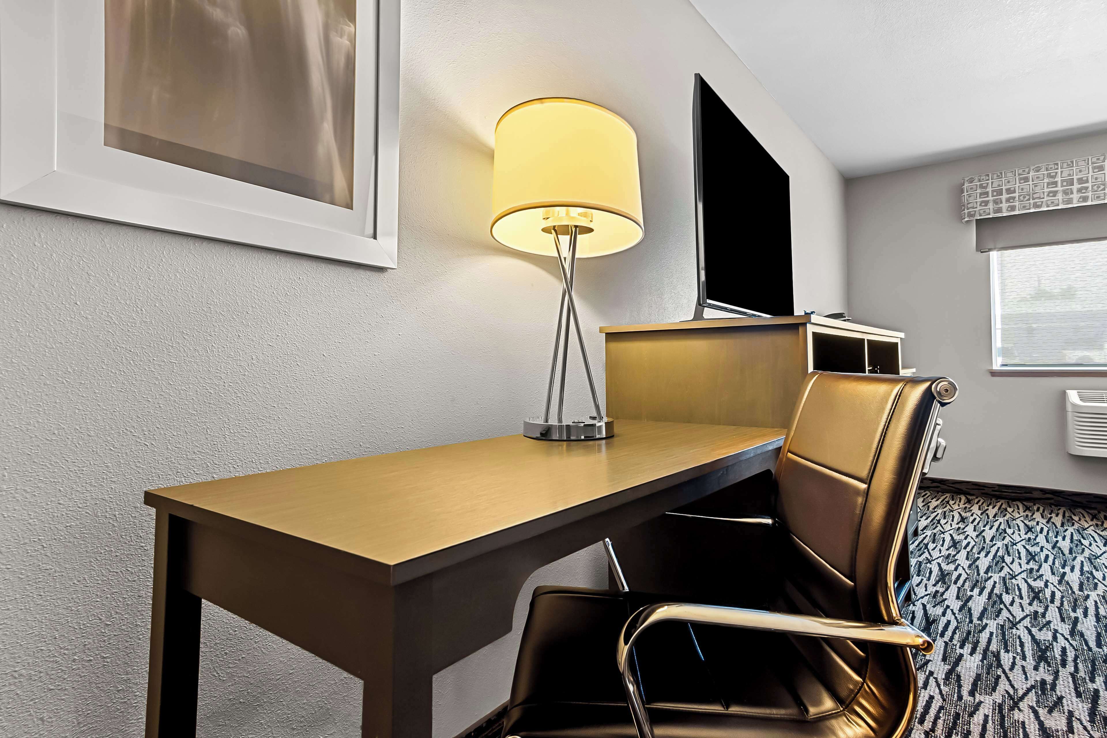 A hotel room with a wooden desk, a black leather chair, a floor lamp, a flat-screen TV, and a patterned carpet.