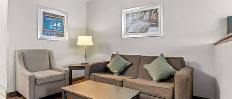 A room with a grey armchair, a brown sofa with two cushions, a small table, and two framed pictures on the wall.