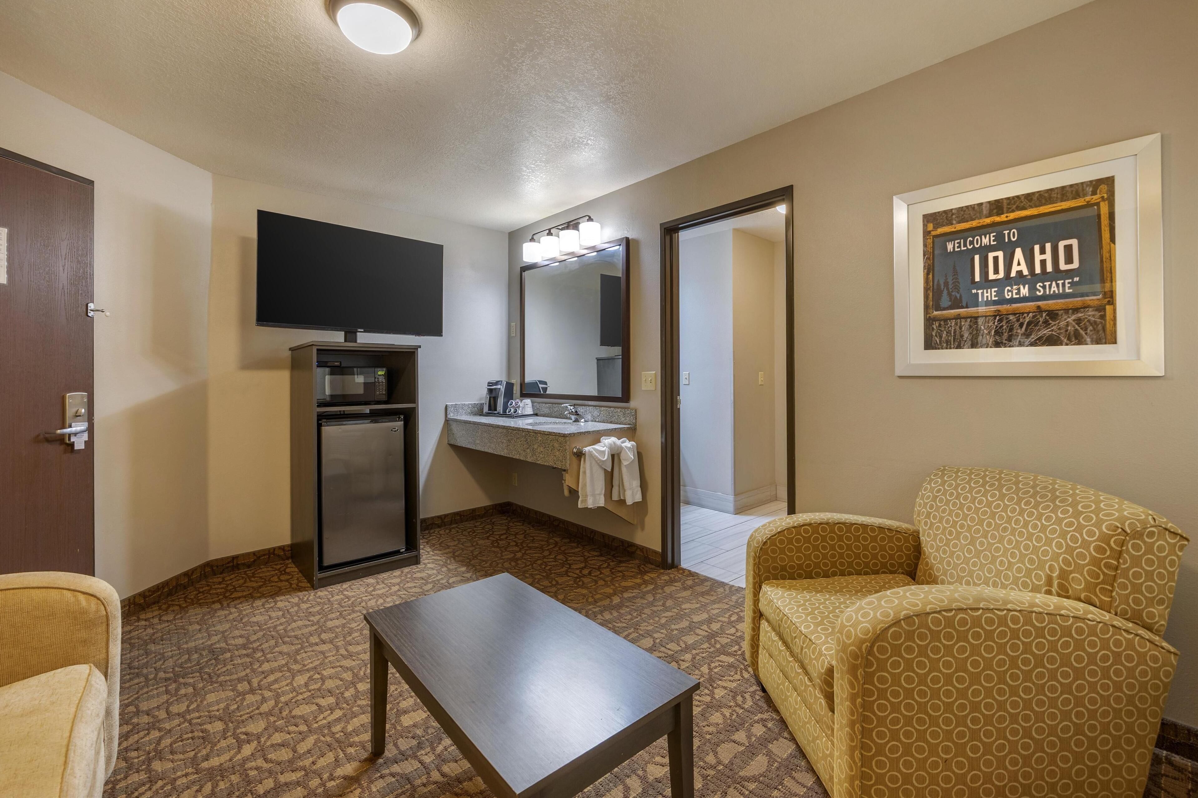 Une chambre d’hôtel équipée d’une kitchenette, d’un téléviseur à écran plat, d’un grand miroir, d’un panneau d’accueil encadré, d’un canapé à motifs et d’une petite table.