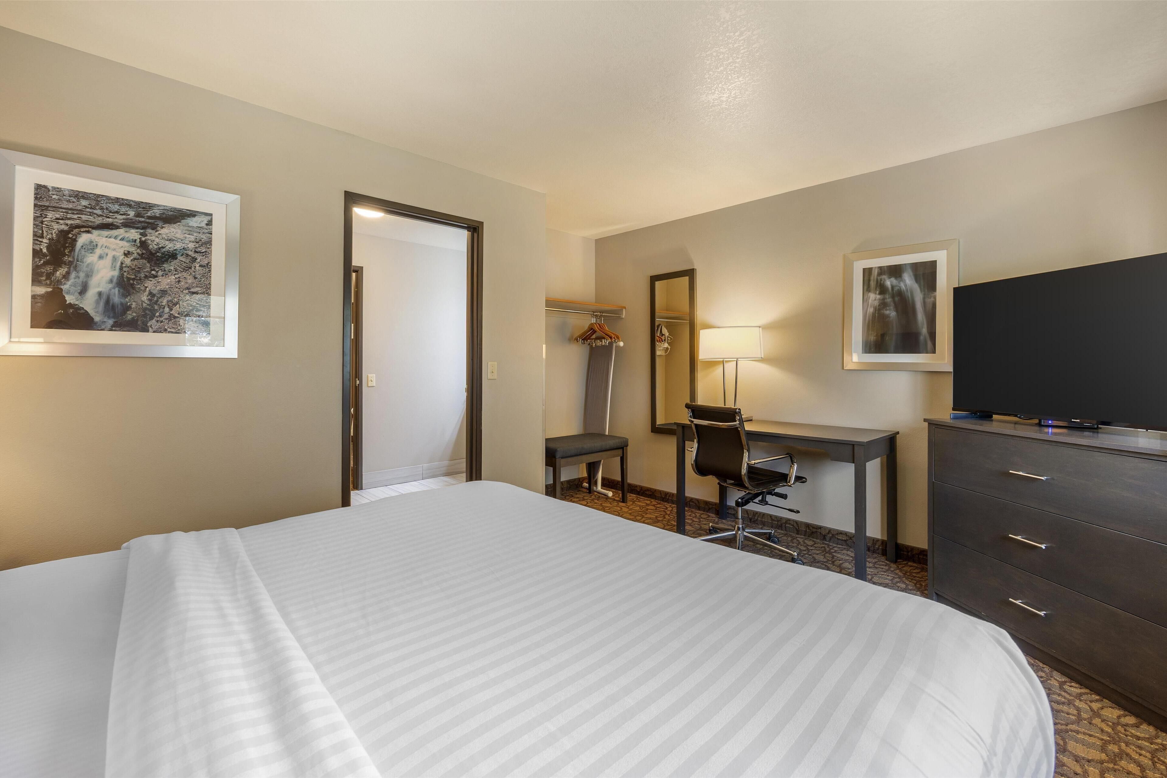 A hotel room with a large bed, a desk with a chair, a dresser, a TV, and two framed pictures on the walls.
