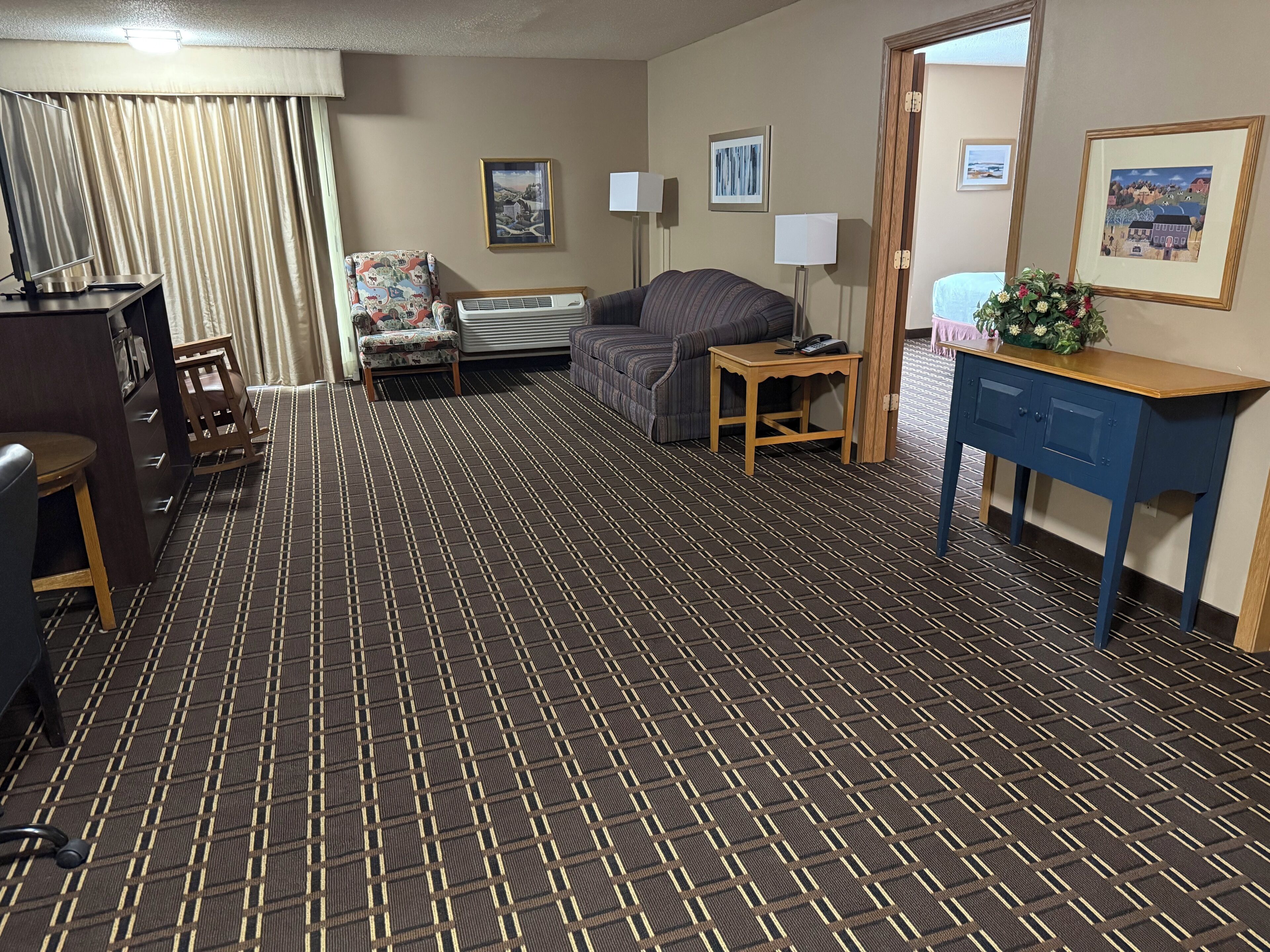 A hotel room with a checkered carpet, a sofa, a desk with a lamp, and a bed.