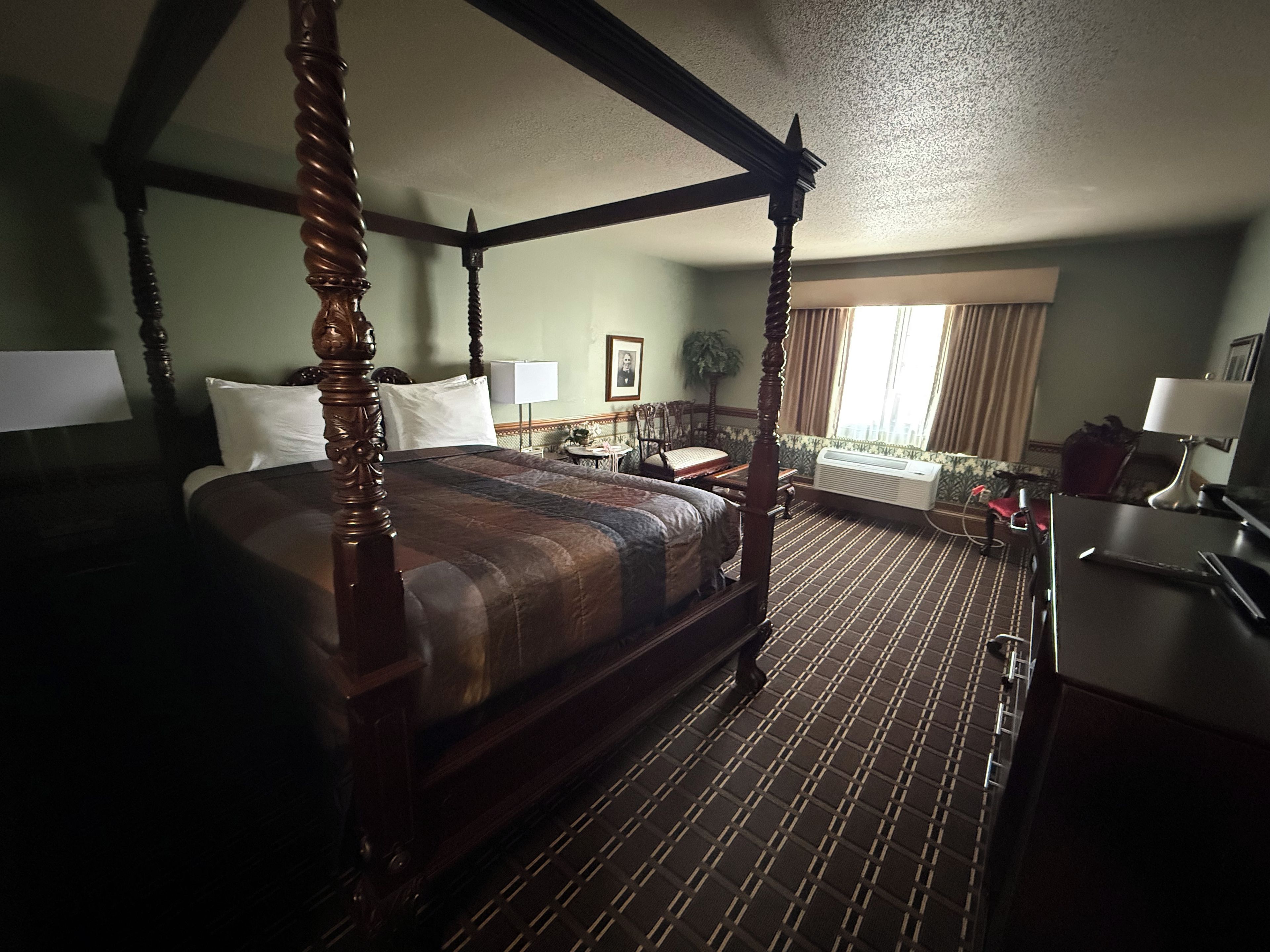 A four-poster bed with a wooden frame and a dark, striped bedspread. There is a desk with a lamp and a chair to the right. The room has a patterned carpet and a window with curtains.