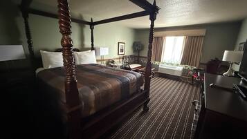 A four-poster bed with a wooden frame and a dark, striped bedspread. There is a desk with a lamp and a chair to the right. The room has a patterned carpet and a window with curtains.