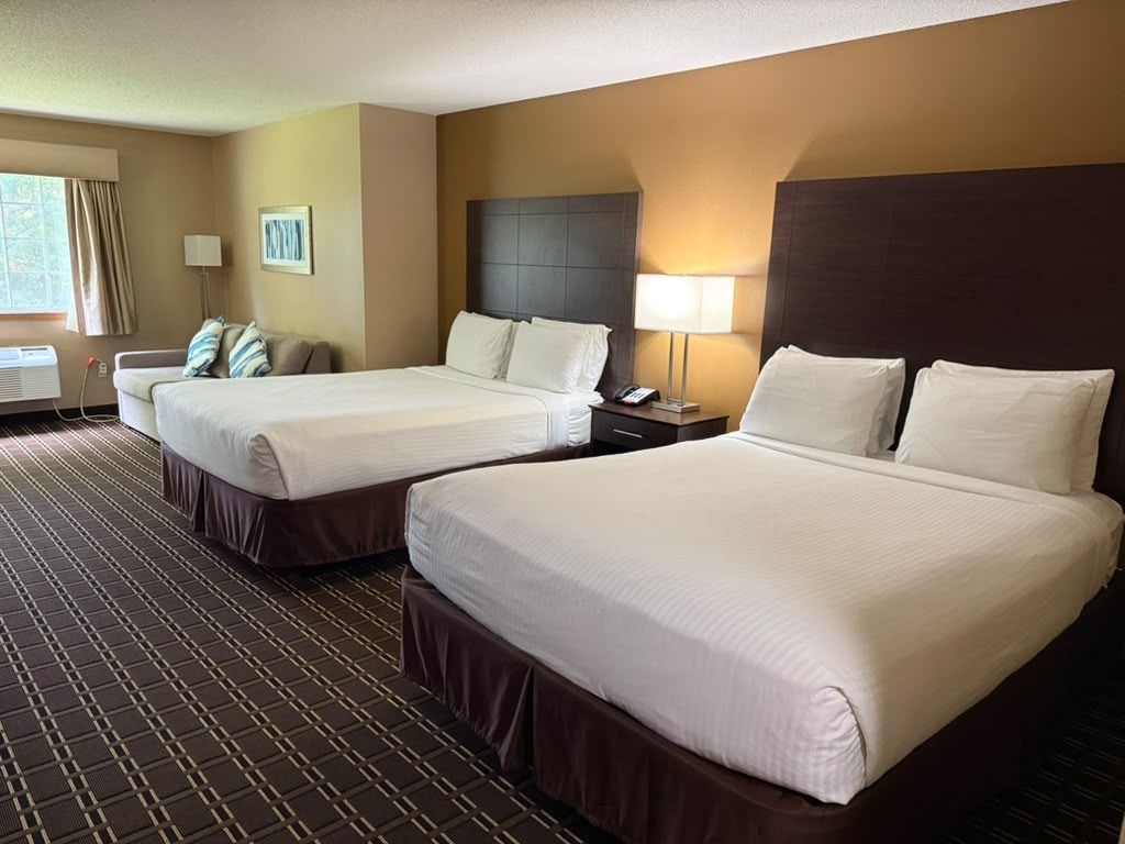 A hotel room with two beds, a headboard, bedside tables, a lamp, and a window with curtains.