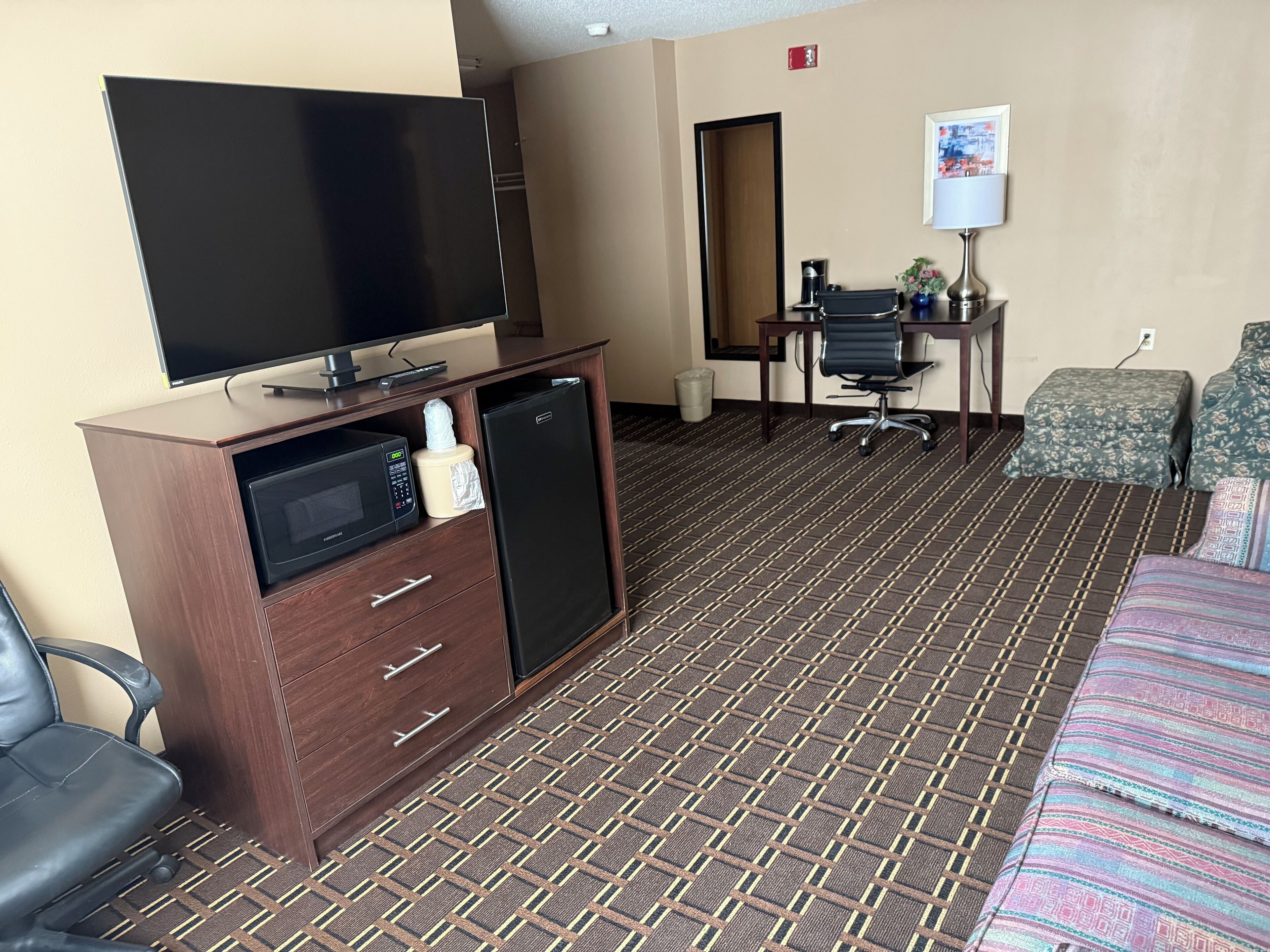A hotel room with a flat-screen TV, a mini-fridge, a microwave, a desk with a chair, a coffee maker, and a bed with patterned bedding.