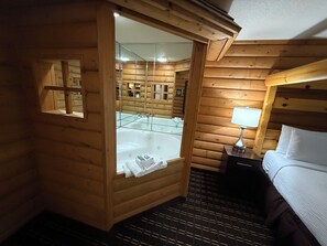 Una habitación de cabaña de madera con una bañera acristalada, una mesita de noche con lámpara y una cama con ropa blanca.