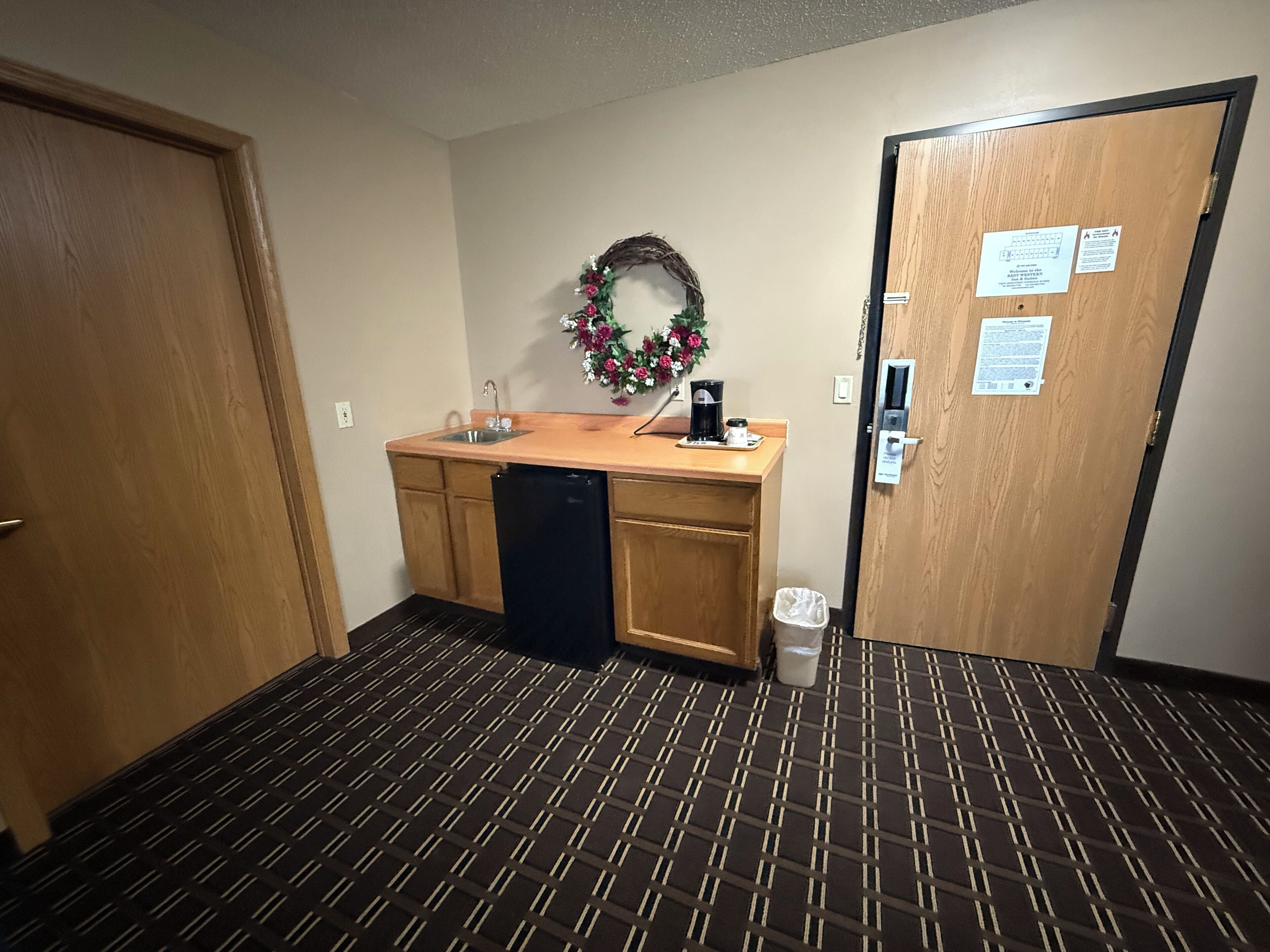 A small kitchenette with a sink, a coffee maker, and a trash can. There is a door with a lock and a sign on it.