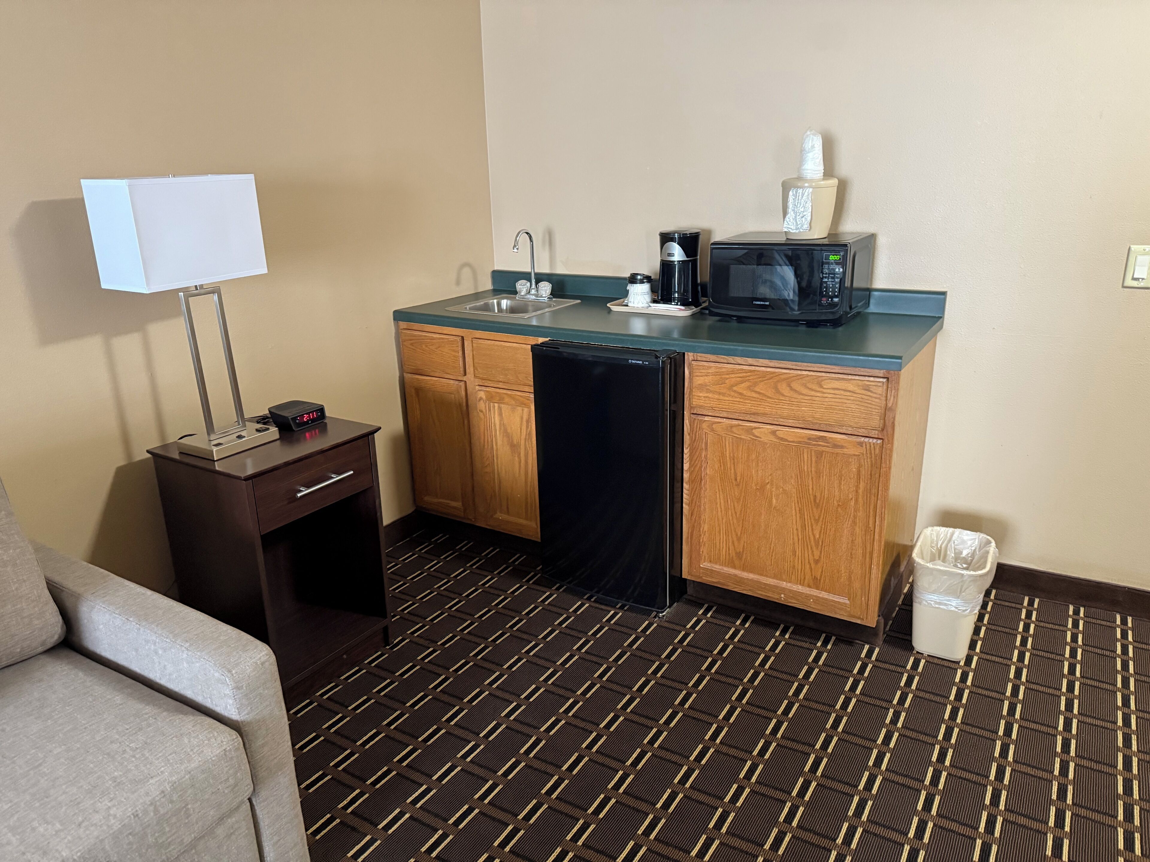 A compact hotel room with a kitchenette, including a microwave, coffee maker, and a small refrigerator. There is a desk with a lamp and a trash bin.