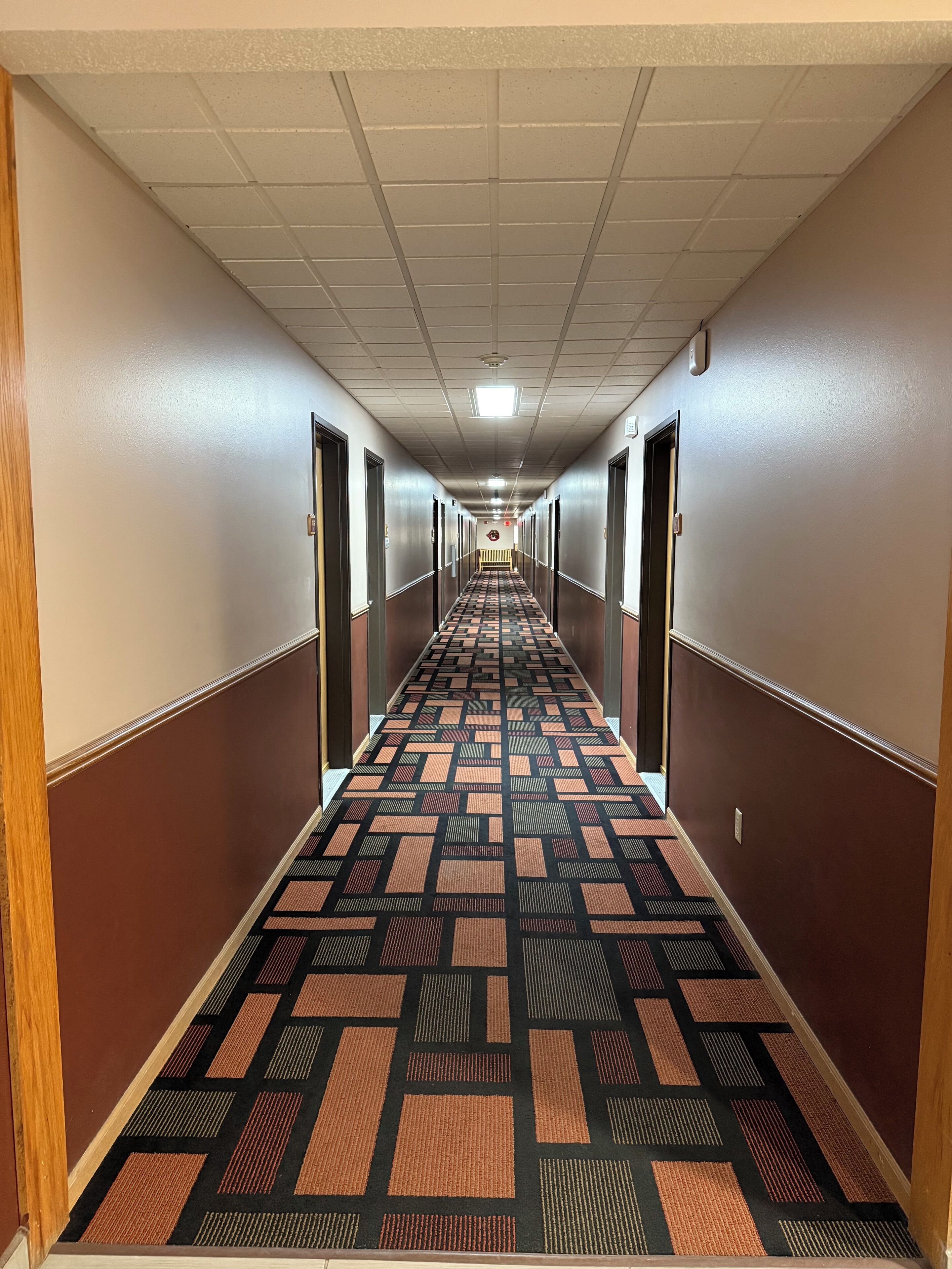 Un long couloir étroit, doté d’un tapis à motifs, avec des portes de part et d’autre, et un plafond éclairé par des néons.