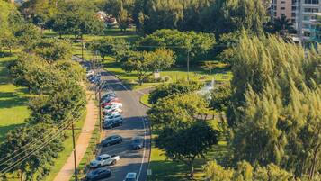 A street lined with parked cars and trees, leading to a park with a fountain.
