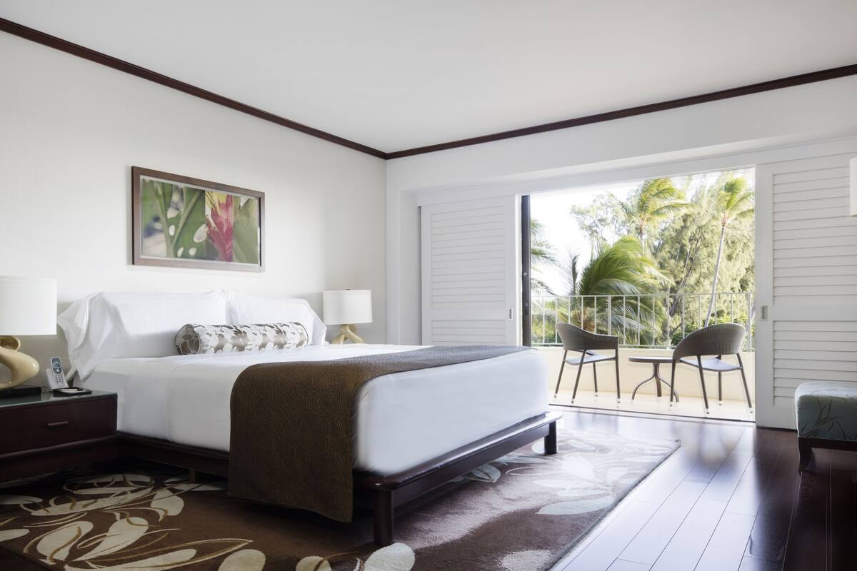 A hotel room with a bed, bedside lamps, a painting, a balcony with chairs, and a view of palm trees.
