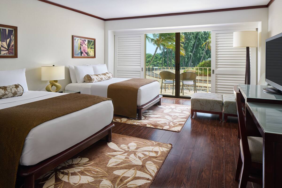 A hotel room with two beds, a desk, a TV, and a balcony with a view of palm trees.